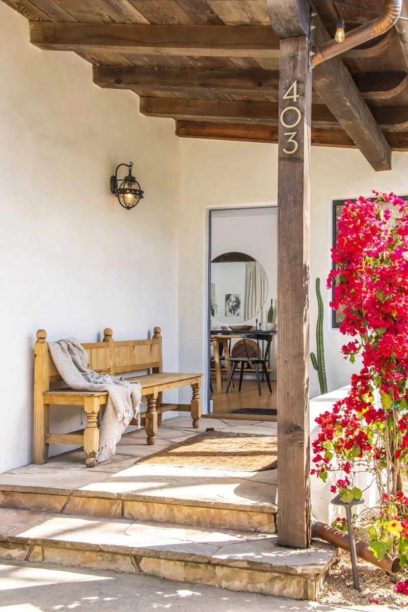 A porch with a wooden bench, a potted cactus, and vibrant pink flowers, leading to an interior room visible through an open doorway.