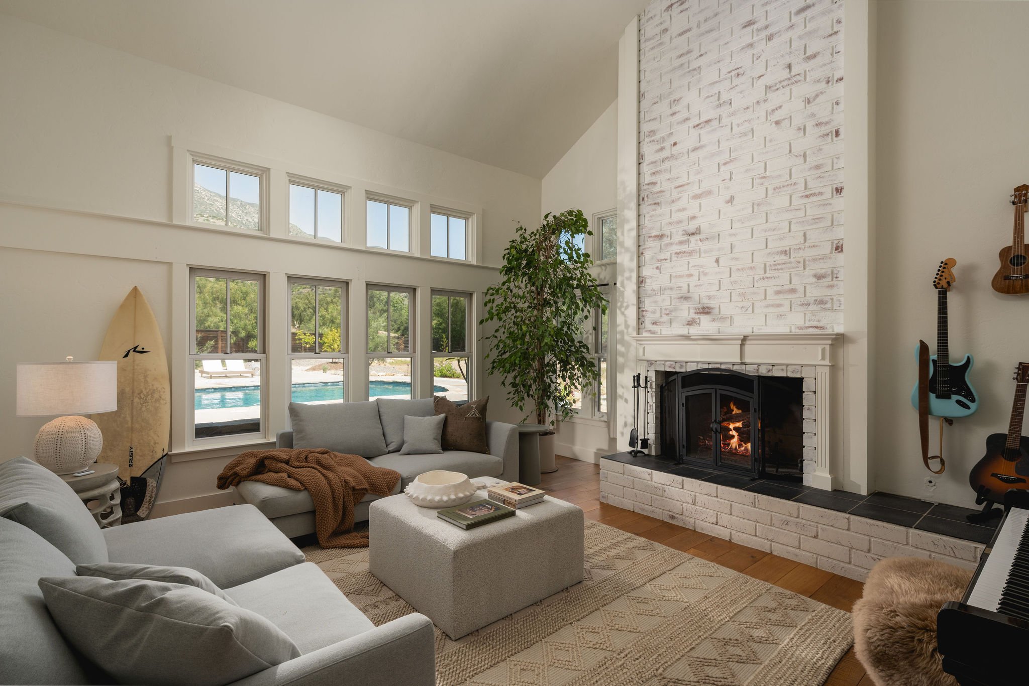 Living room with large windowed wall overlooking a pool and mountain view, white brick fireplace, gray sofas, plants, guitars, and a keyboard.