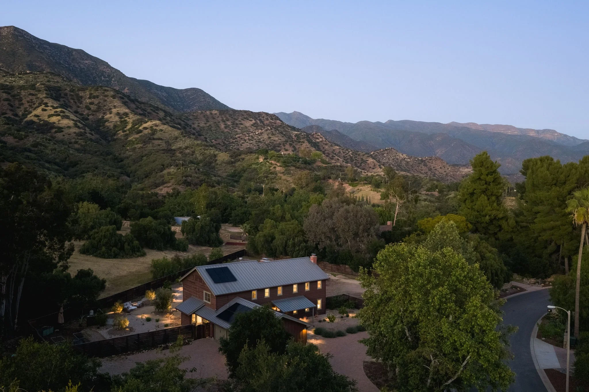 A view of a mountainous landscape with houses in the foreground during dusk. The sky is clear and the houses have lights on, with some solar panels visible on the roof.