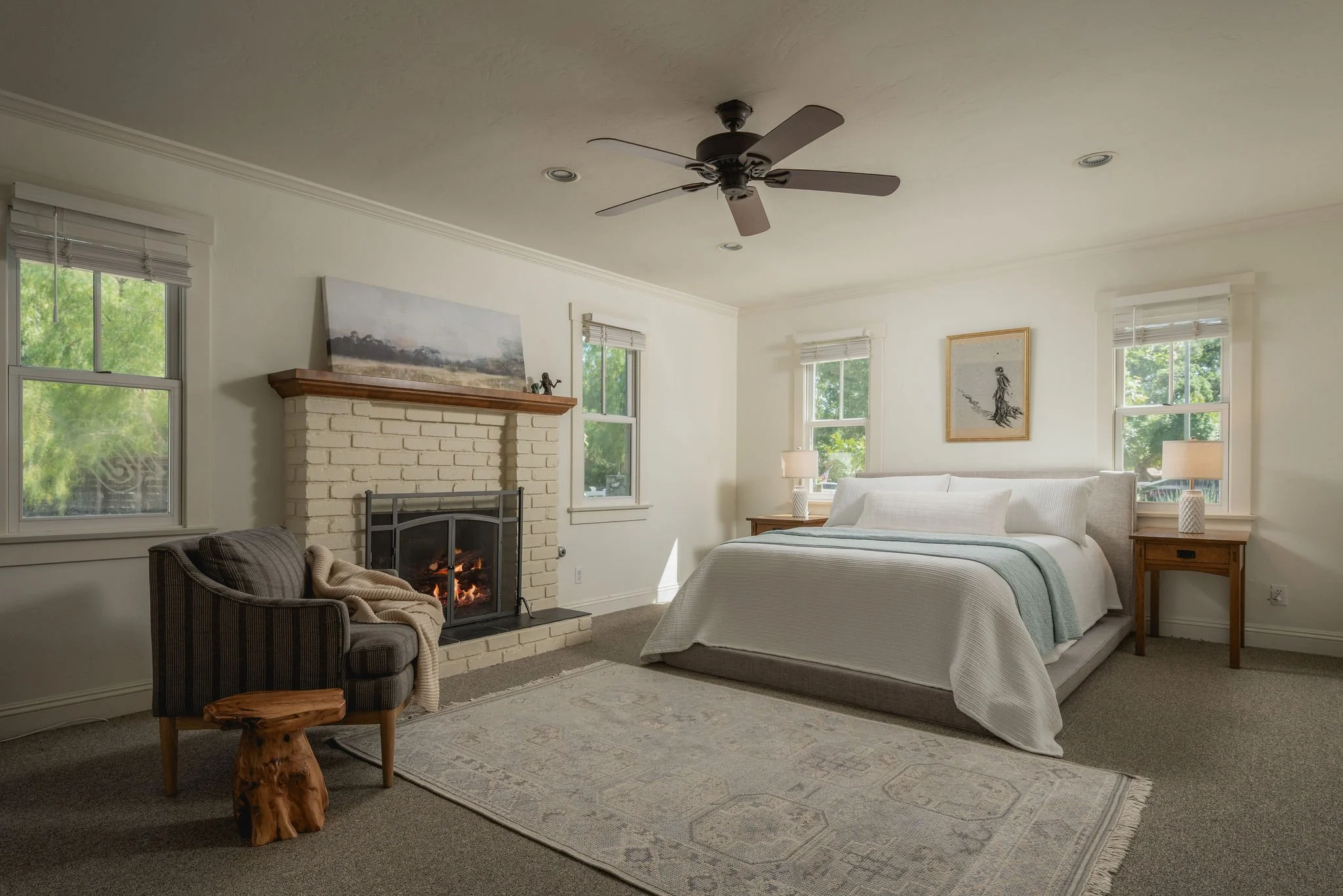 A bedroom with a fireplace, a bed, two bedside tables with lamps, a chair, a small table, and artwork on the walls.