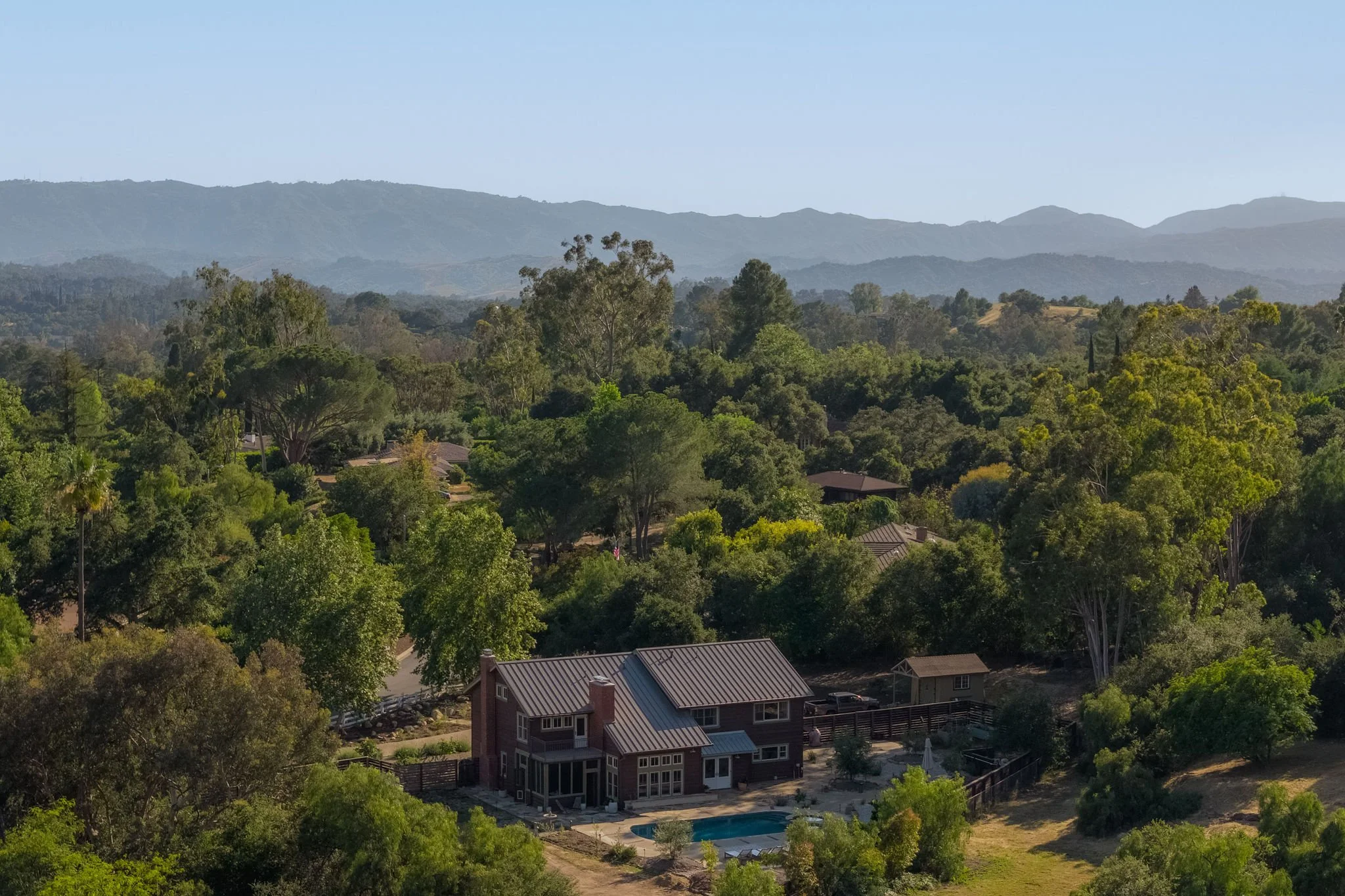 A scenic view of rolling mountains in the distance with a lush green forest in the foreground, a residential house with a backyard swimming pool, and a clear blue sky.