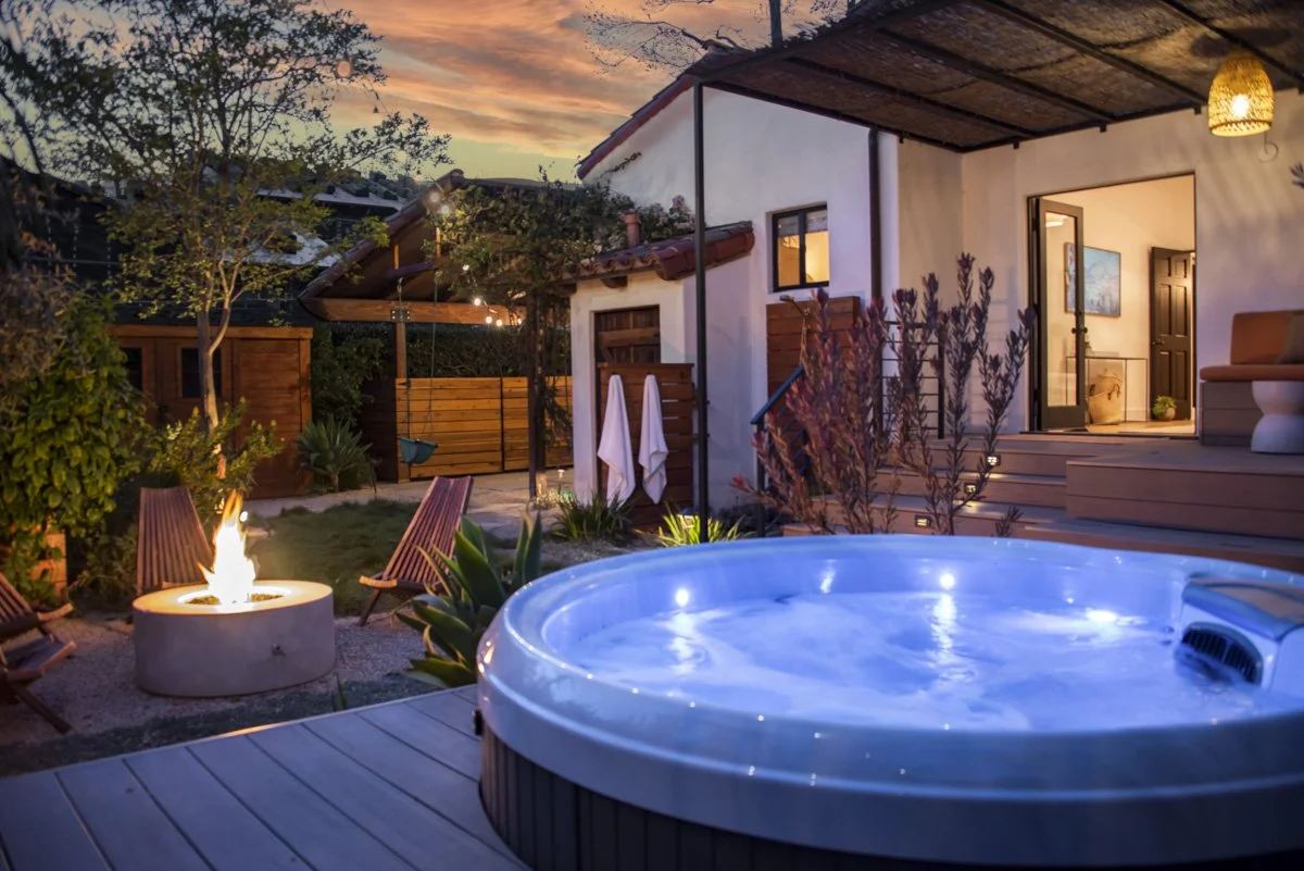Backyard outdoor space at dusk with a hot tub, fire pit with flames, wooden chairs, and a house with steps leading to an open door.