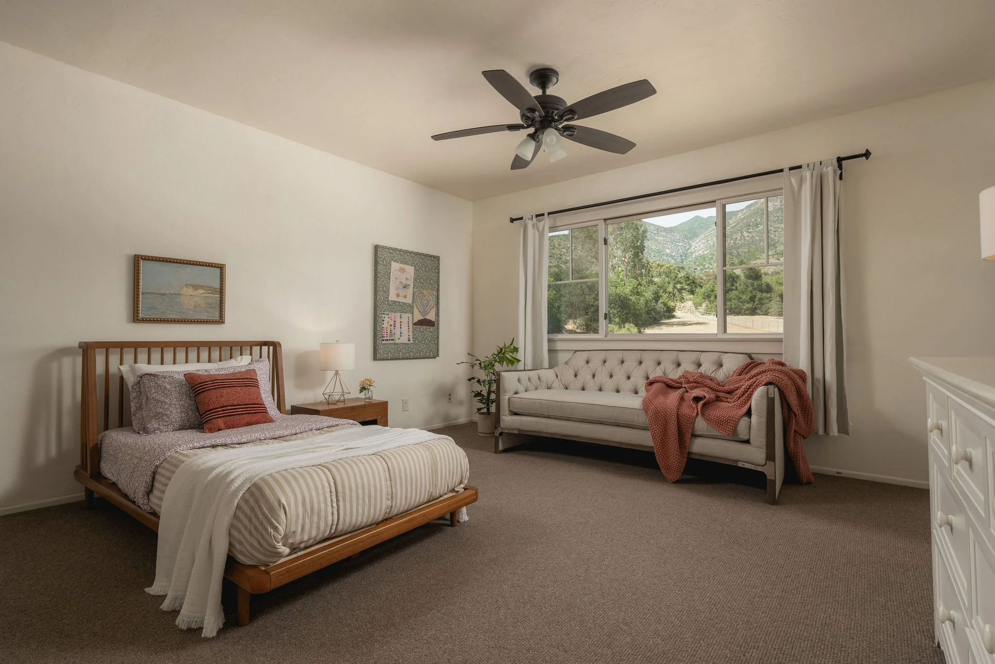 A bedroom with a wooden bed, a nightstand, and a white couch with a blanket and pillows. There is a lamp on the nightstand, artwork on the wall, and a large window with curtains showing a view of green trees and mountains outside.