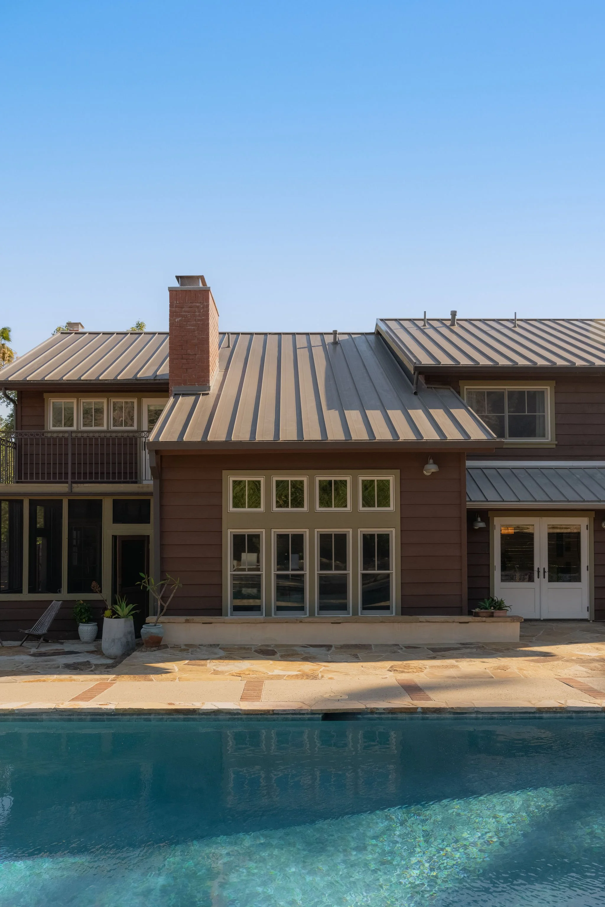 Backyard view of a house with a metal roof and chimney, pool in the foreground, and clear blue sky.