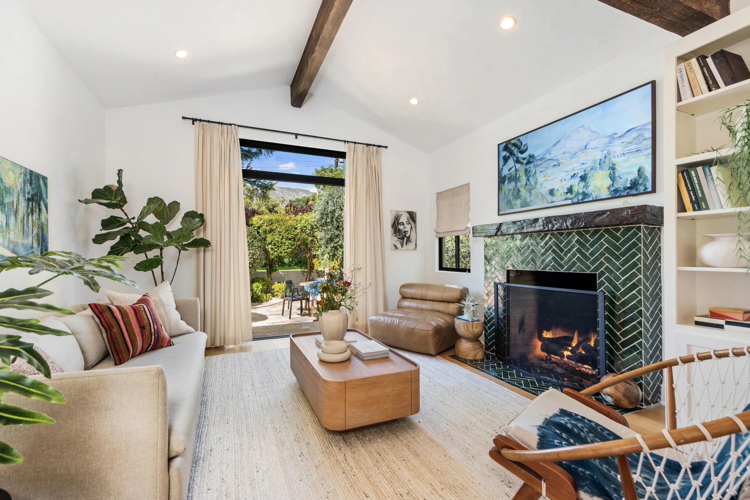 Living room with white walls, beige couch, brown leather armchair, wooden coffee table, large window with curtains, fireplace with green tile, wall art, bookshelves, and indoor plants, with outdoor patio visible through window.
