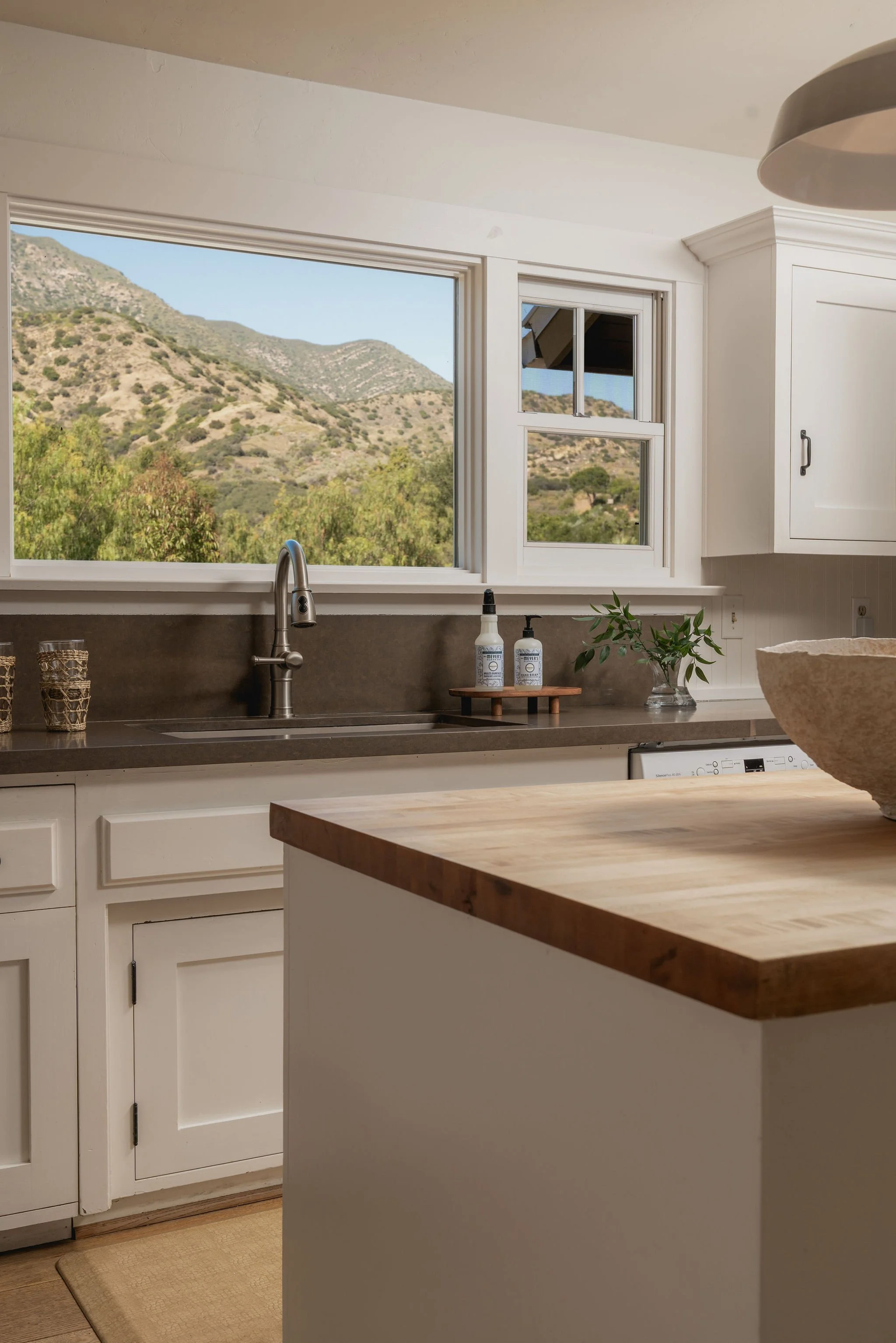 Kitchen view with a large mountain landscape outside the window, white cabinets, a sink with a modern faucet, and various decorative and functional items on the countertop.