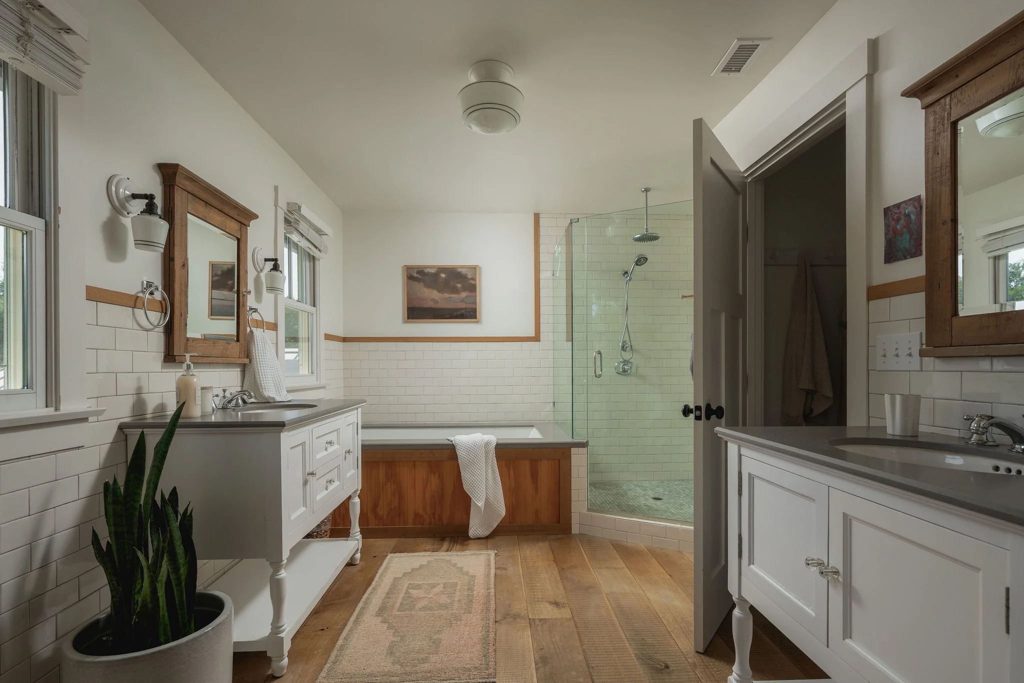 A bathroom with white subway tile walls, wooden accents, a walk-in tiled shower with a glass door, a bathtub, double vanity sinks, framed mirrors, wall-mounted lights, windows with blinds, a framed painting, a potted plant, a beige rug, and wooden fl