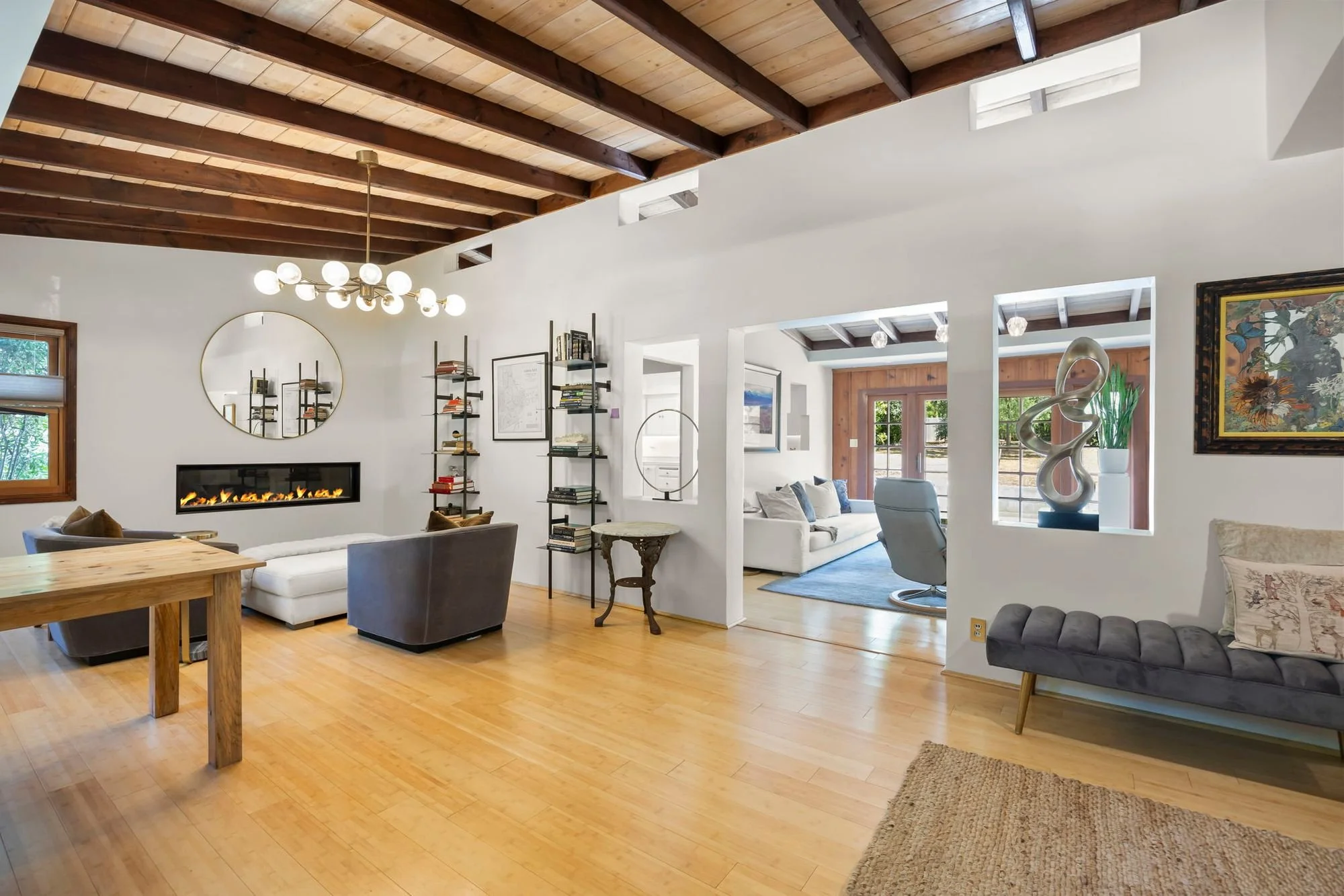 A spacious living room with a wood-paneled ceiling, hardwood floors, modern furniture, and a fireplace. The room features large windows, a decorative sculpture, a bookshelf, and a round mirror on the wall.