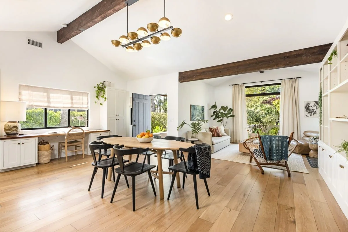 Open living and dining area with wooden floors, white walls, and large windows with curtains. Dining table with six black chairs, living area with cream sofa, armchair, and large windows with greenery outside. Ceiling with exposed wooden beams and mo