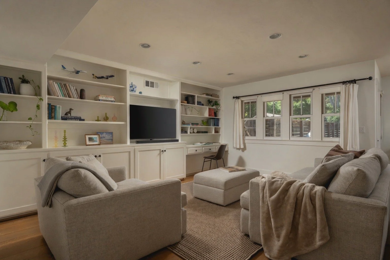 Living room with beige sofa and armchair, built-in white bookshelves, flat-screen TV, window with white curtains, house plants, and wooden floors.