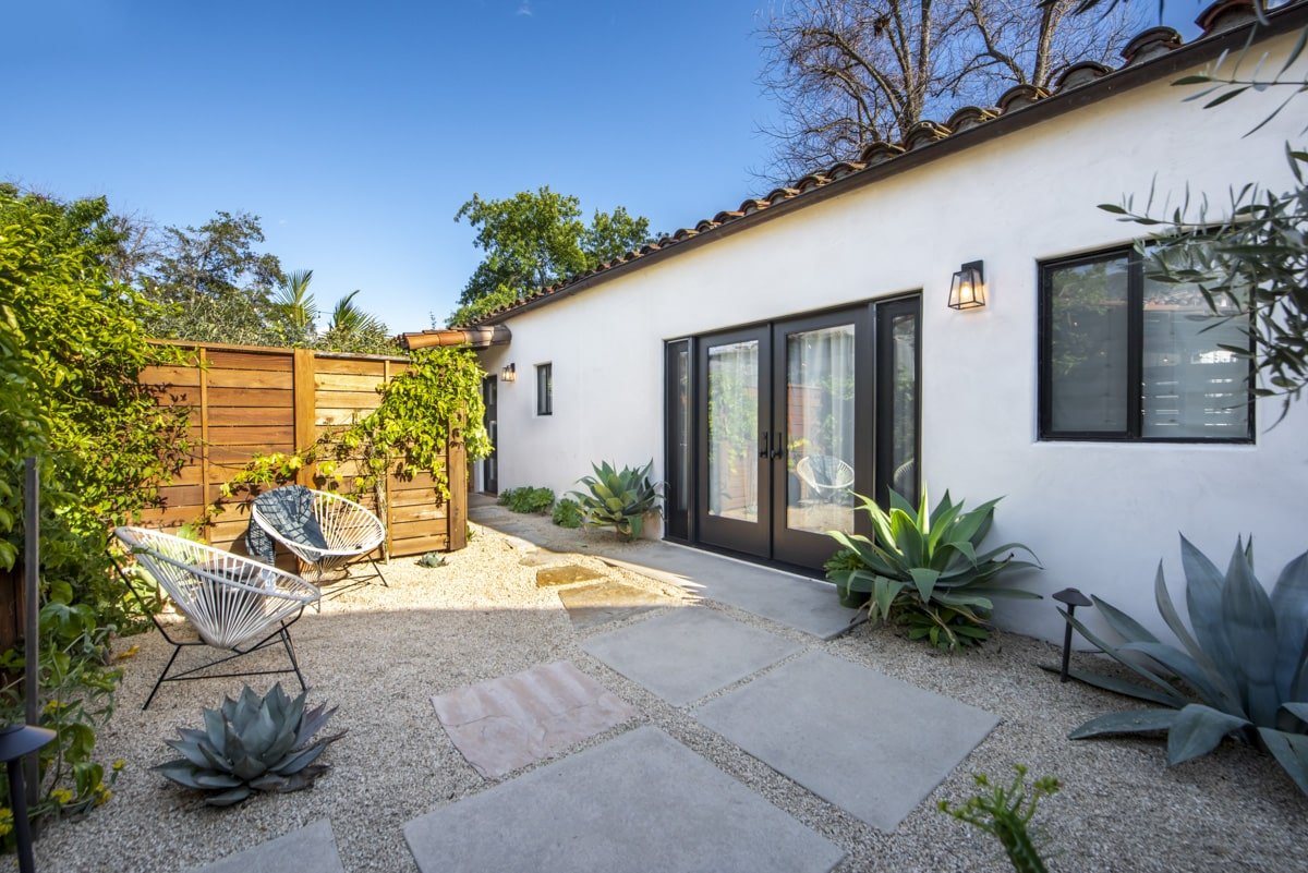 A backyard patio with white gravel and large rectangular stepping stones, featuring outdoor seating with white modern chairs, various plants including succulents, and a white house with black-framed windows and glass doors, surrounded by a wooden pri