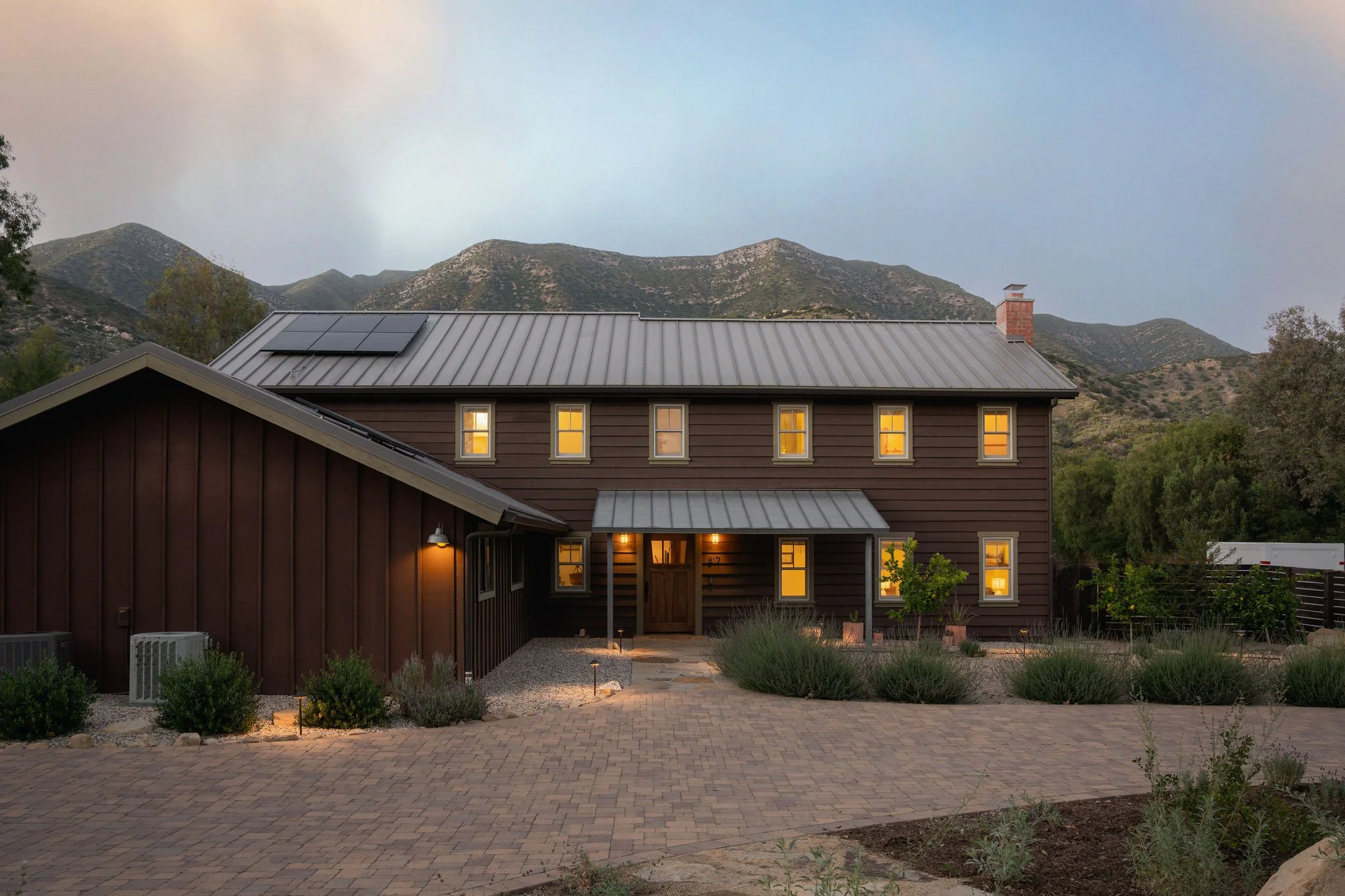 A house with dark brown exterior walls and a metal roof, illuminated windows, set against a backdrop of green mountains under a cloudy sky at dusk.