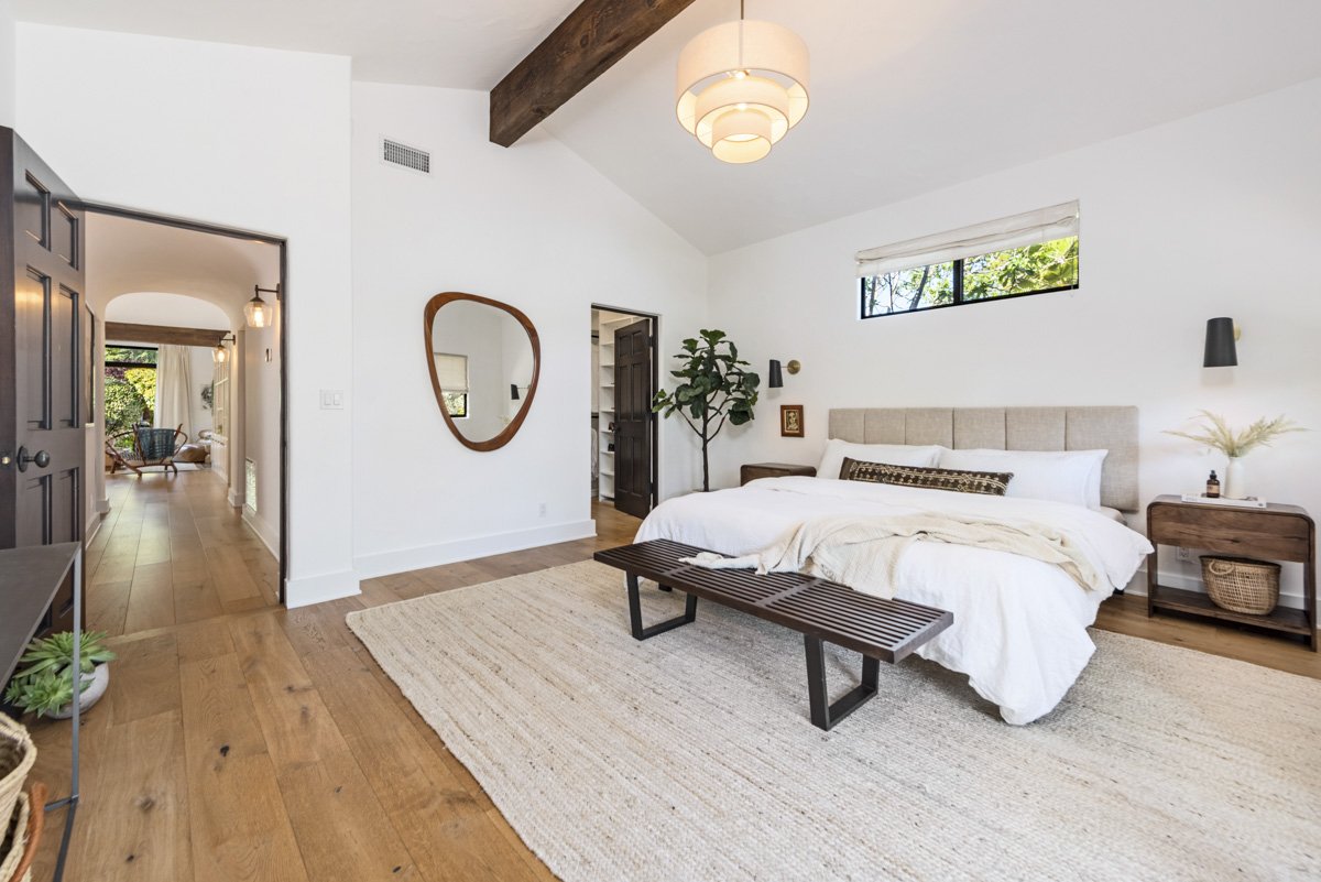 A modern bedroom with white walls, wooden floors, and a high ceiling with a wooden beam. There's a bed with a beige headboard, white bedding, and a decorative pillow. A wooden bench sits at the foot of the bed. To the right, there's a nightstand with