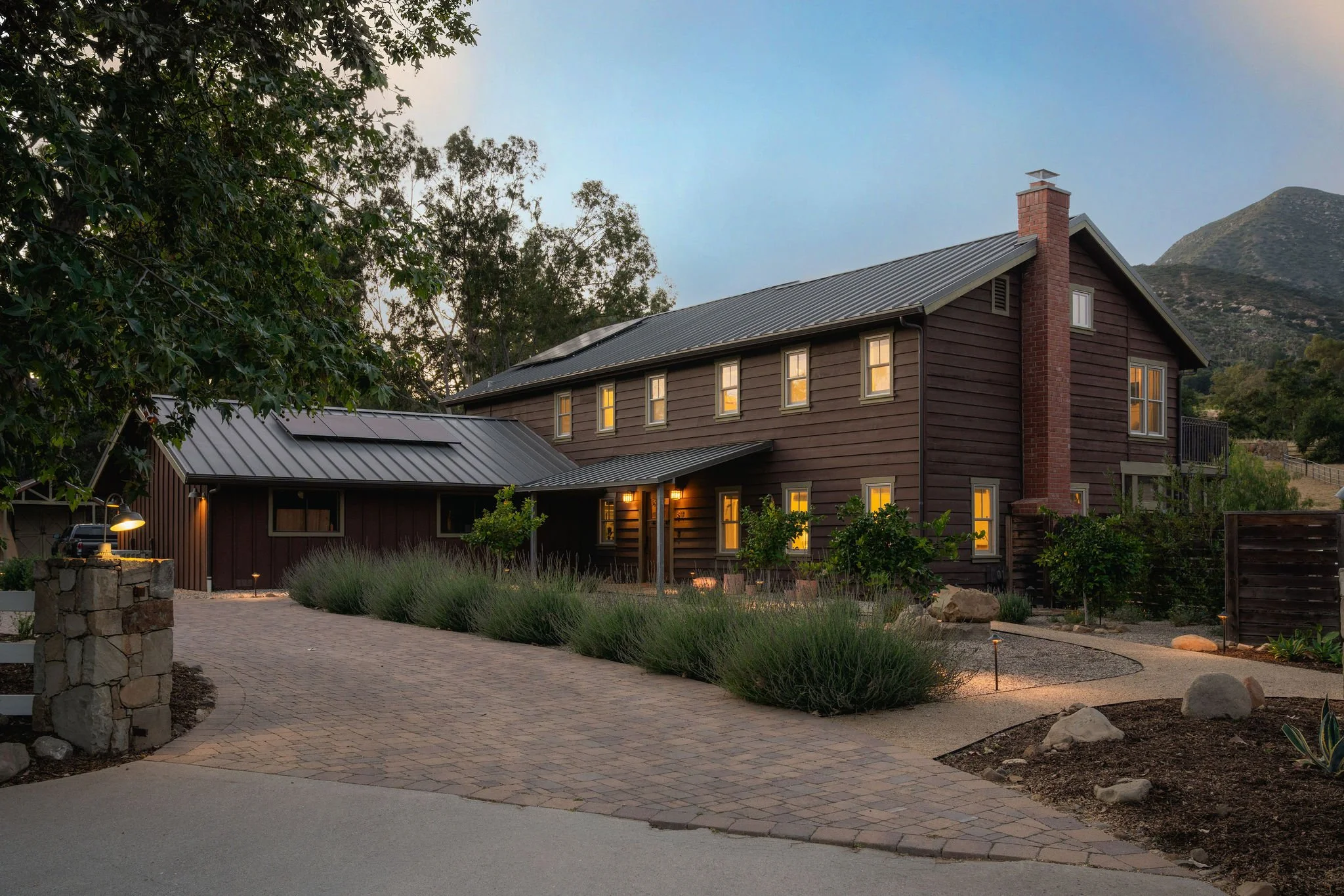 A two-story wooden house with a metal roof, lit windows, a brick chimney, surrounded by trees and landscaped plants, with a mountain in the background and sunset lighting.