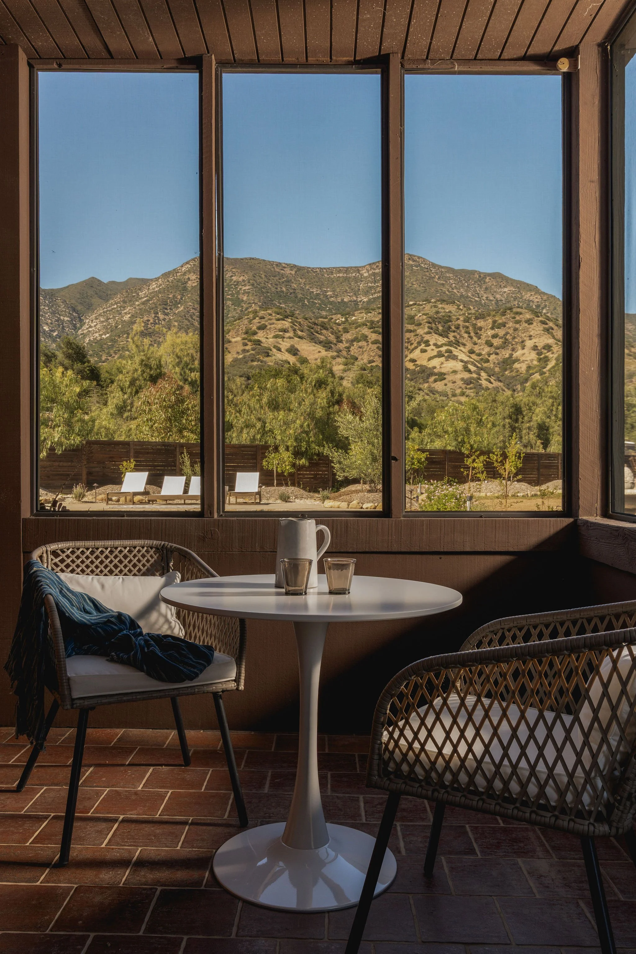 A cozy indoor space with a round white table, two wicker chairs with cushions, and a view through large windows of green hills and trees in the background.