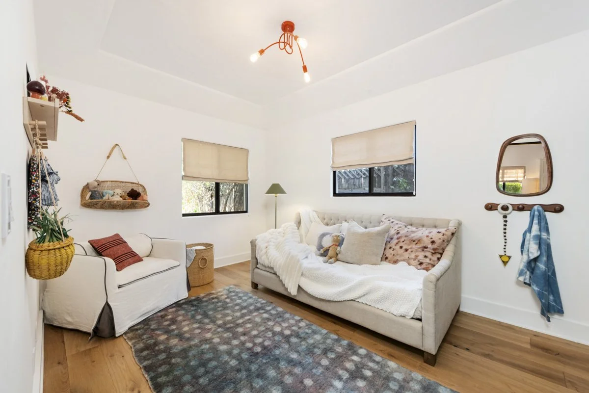 A cozy living room with white walls and wooden flooring, featuring two windows with beige blinds, a beige sofa with pillows and a stuffed animal, a white loveseat with a pillow and a blanket, a patterned area rug, a green floor lamp, a wall mirror, a