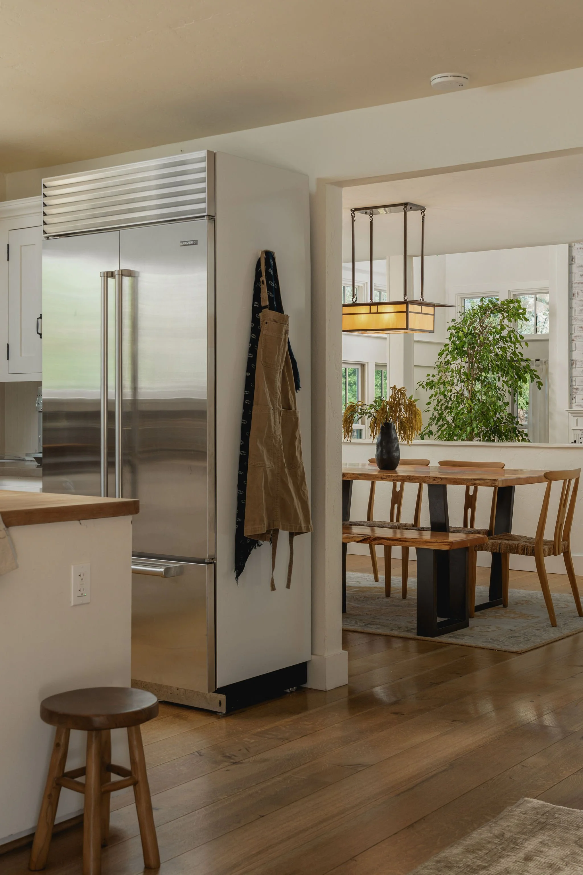 View of a modern kitchen and dining area with wooden floors, a stainless steel refrigerator, a round wooden stool, a dining table with chairs, a large plant, and a hanging light fixture.