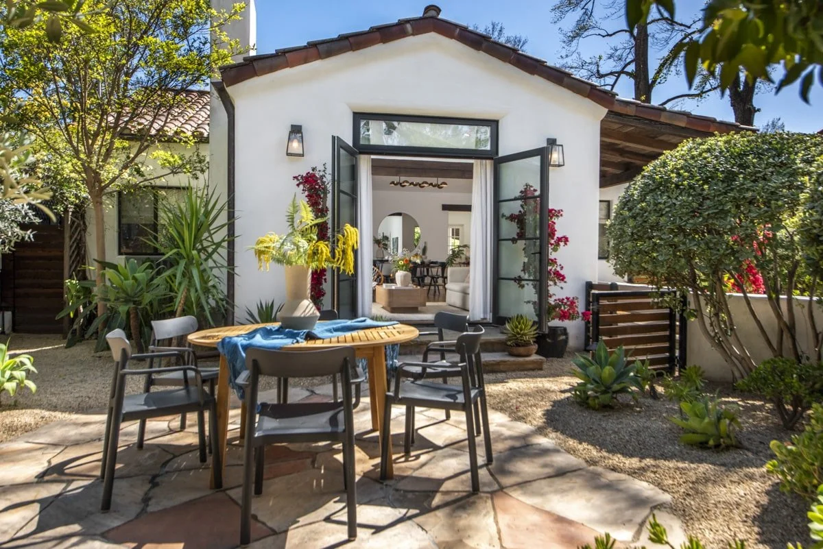 Exterior view of a house with open glass doors leading to a living room, surrounded by a garden with various plants and trees, outdoor furniture, and a stone patio.