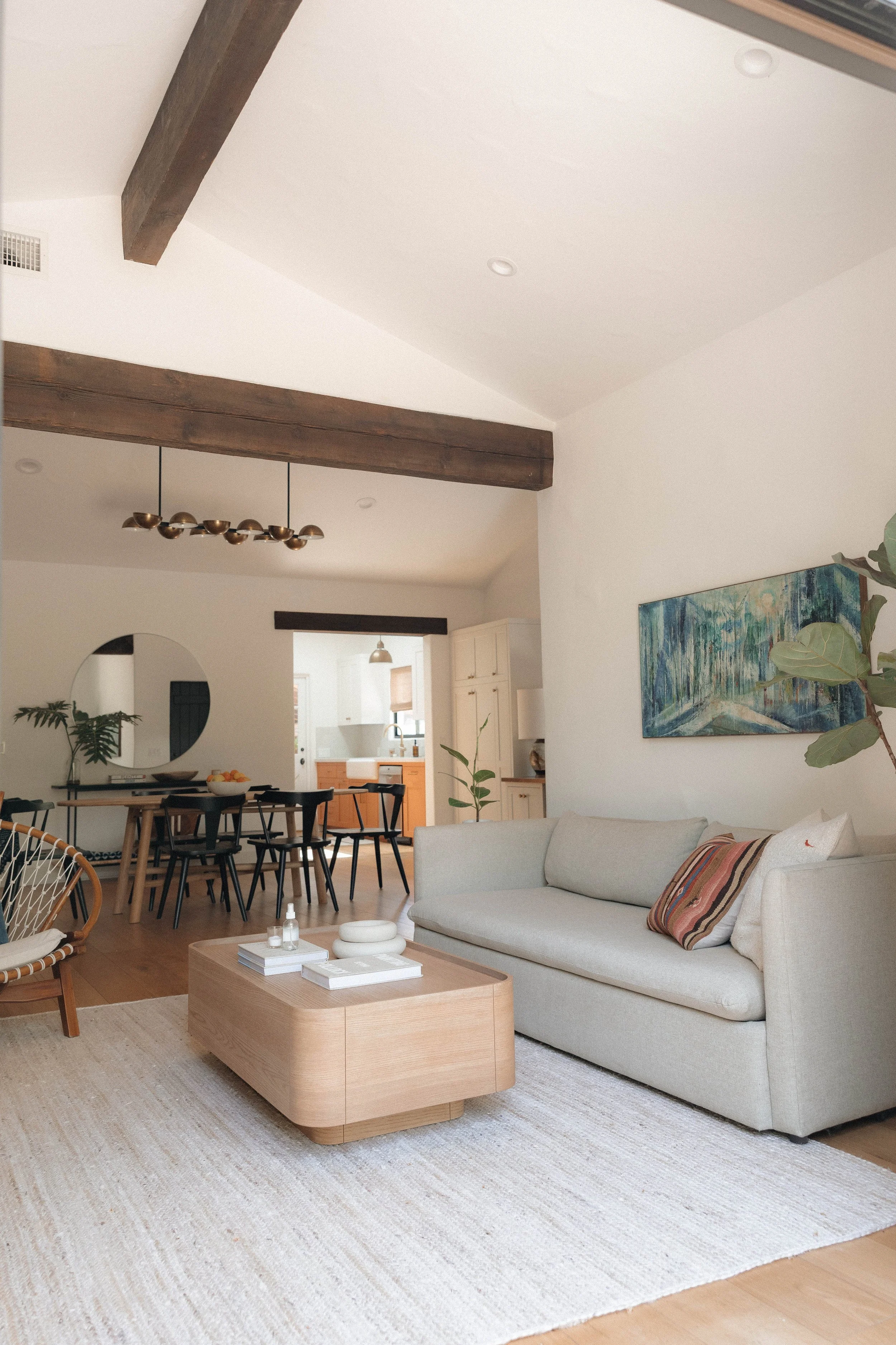 Living room with a white sofa and a wooden coffee table, decorated with books and vases, on a cream-colored rug. A large painting of a forest scene hangs on the wall, and there are houseplants. The open-concept space includes a dining area with black