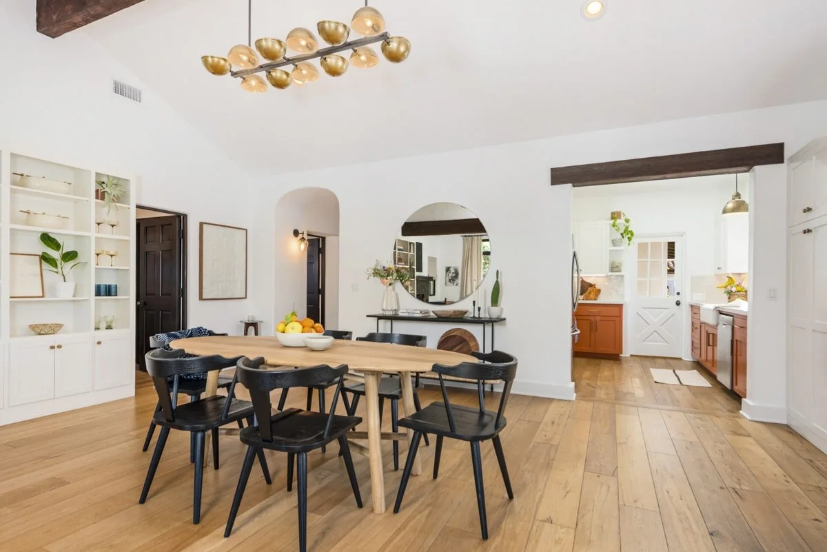 Open-concept dining and kitchen area with a round dining table, black chairs, light wood floors, white walls, decorative shelves, and modern lighting fixtures.