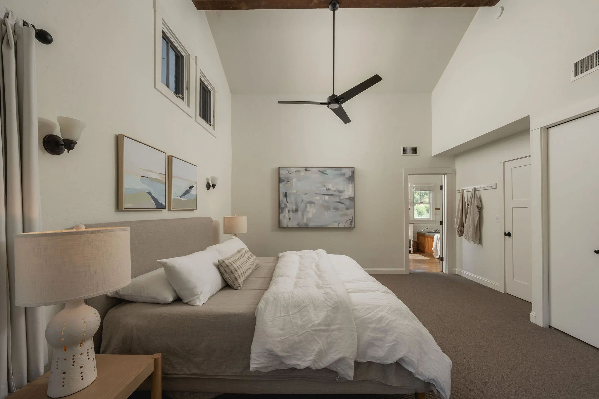 A bedroom with a bed, two table lamps, wall art, windows, a ceiling fan, and a door to an adjacent room.