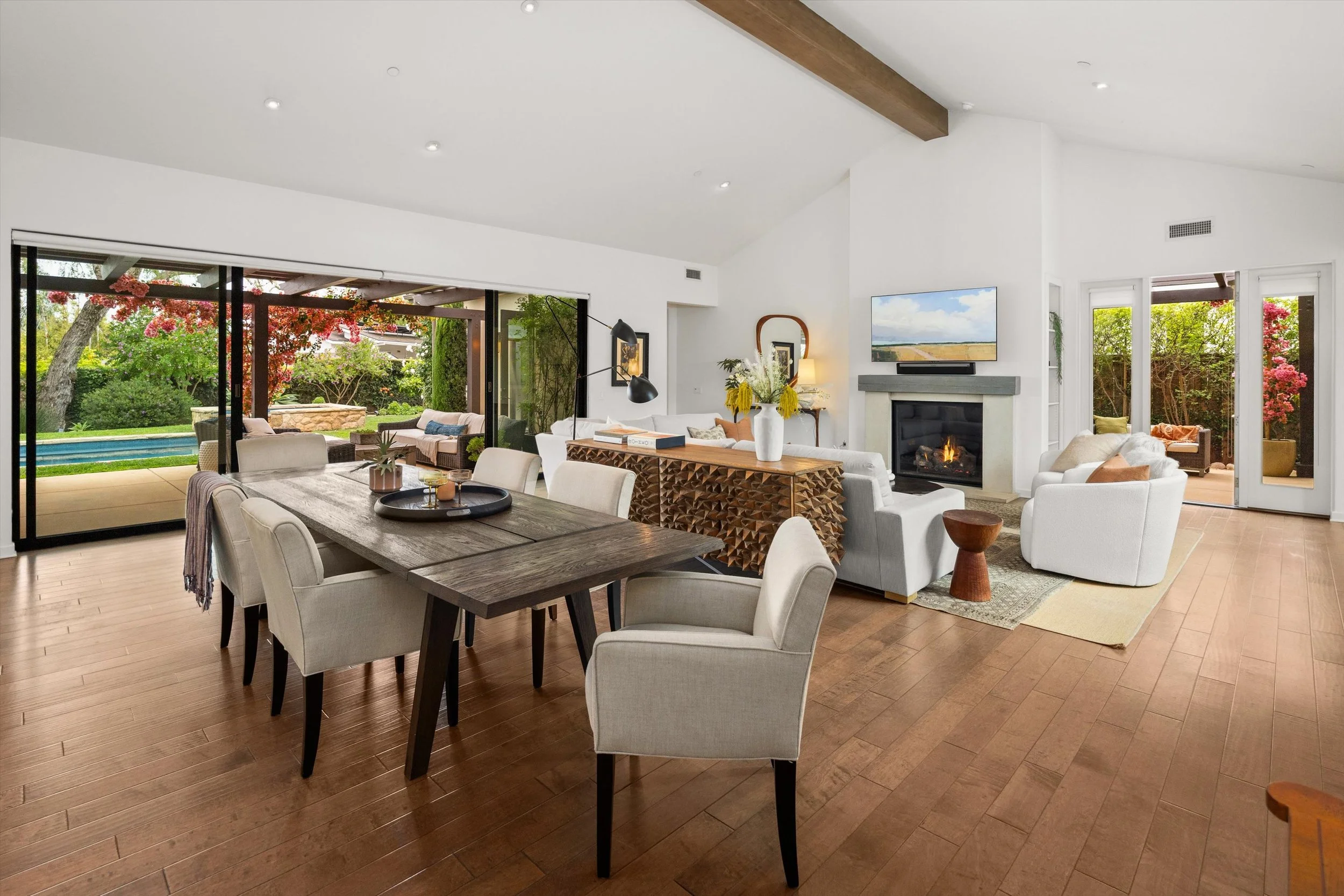 Modern living room with a dining table, white chairs, and a lounge area with white sofas. Large sliding glass doors open to a backyard with a pool, trees, and flowering bushes. There is a television mounted above a fireplace on a white wall, and various decorative items including a large vase with flowers on a wooden sideboard.