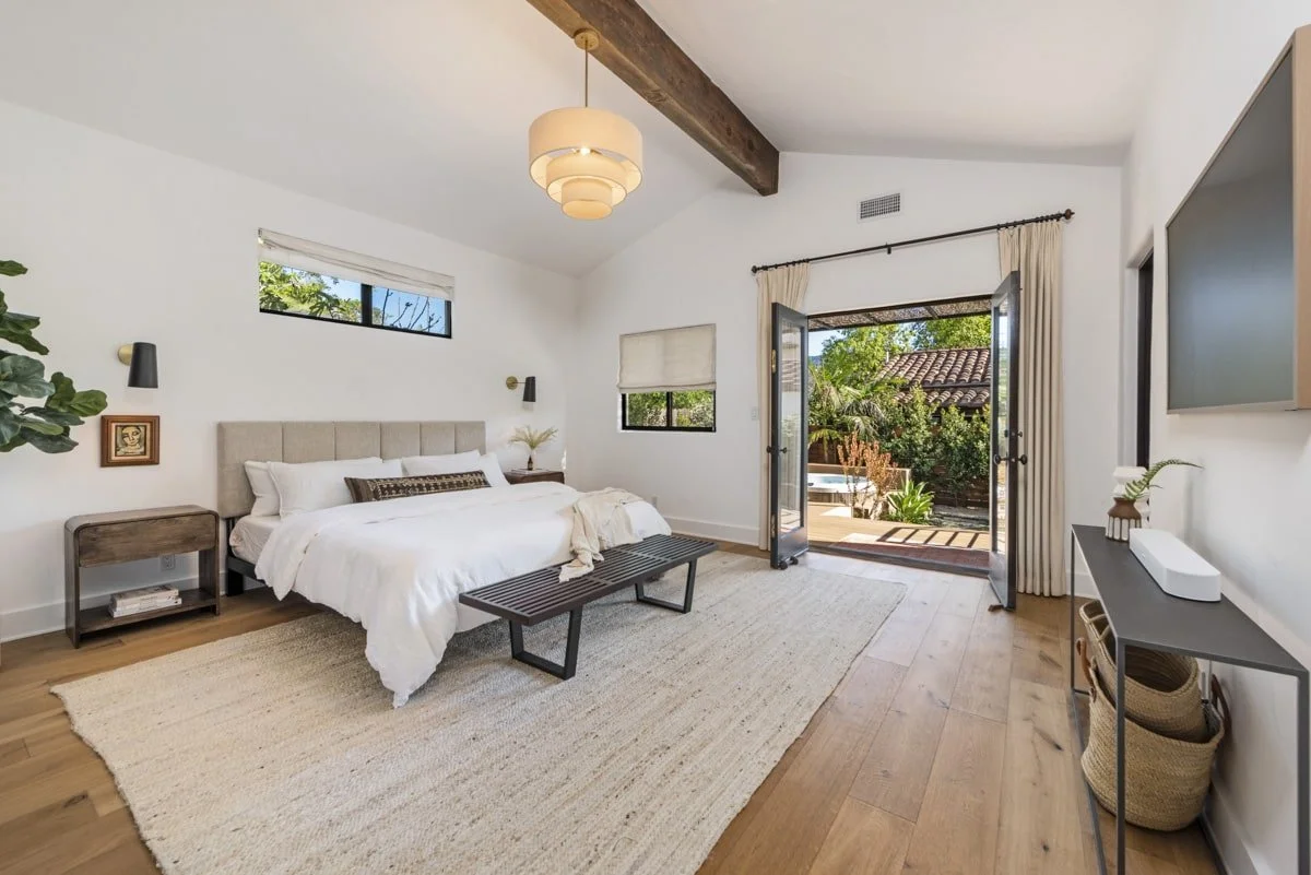 A bedroom with a white bed, beige headboard, and small nightstands, one with books. There is a bench at the foot of the bed. The room has white walls, wooden ceiling beams, and hardwood floors. Double glass doors open to a patio with outdoor greenery
