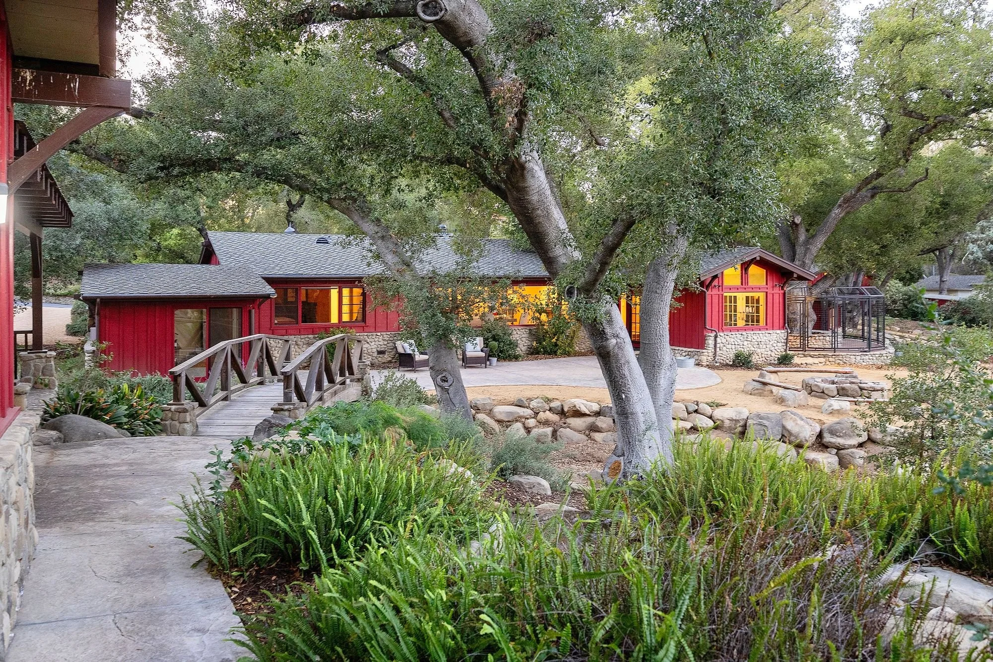 A backyard with large trees, a stone and wooden pathway, and red houses with yellow-lit windows. There are green plants, a stone fire pit, and benches in the yard.