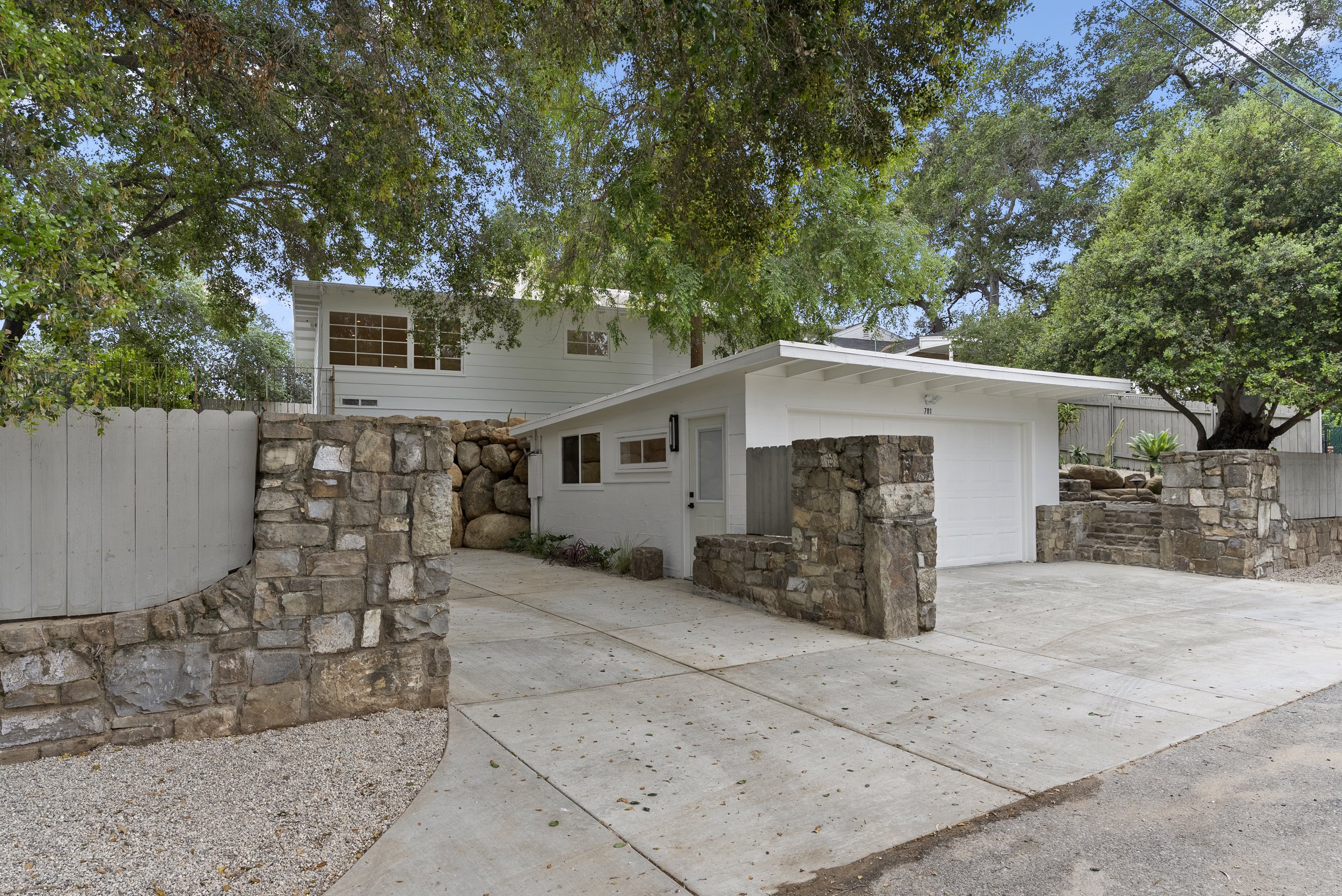 A modern house with a white exterior and large windows, surrounded by trees, with a stone and wood fence in the front yard, and a driveway leading up to a garage.