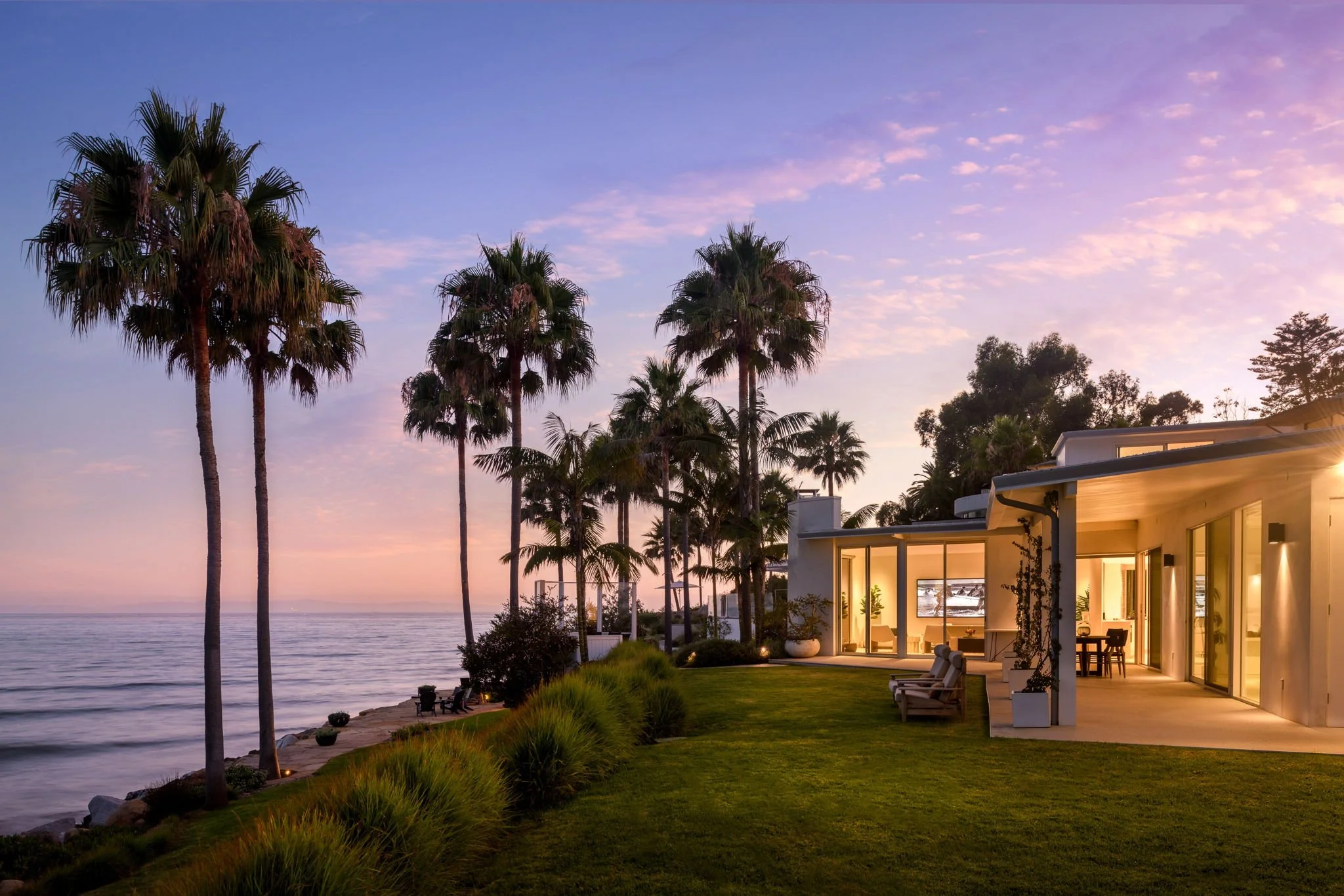 Modern house by the water with tall palm trees, lush green lawn, and a sunset sky with pink and purple hues.