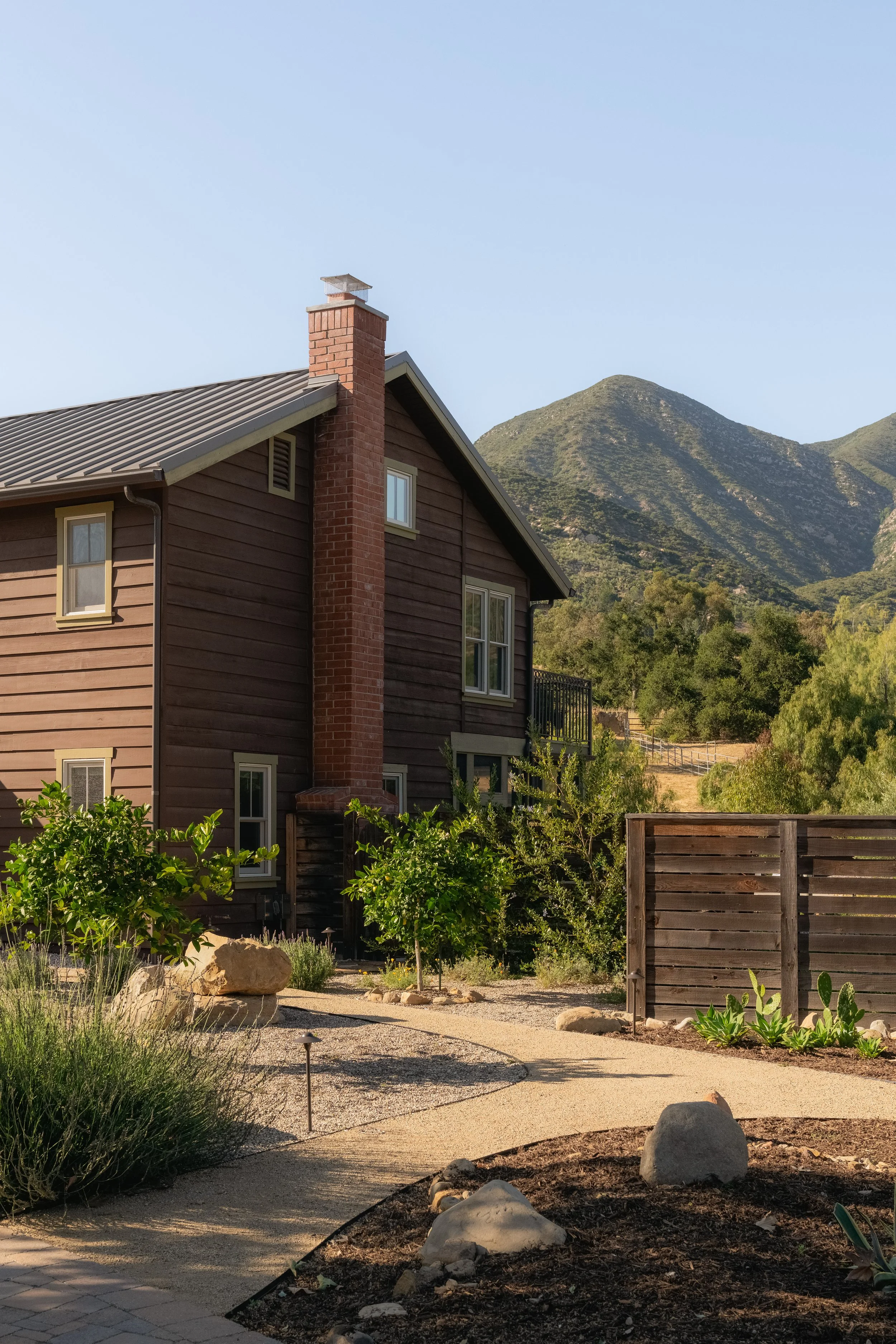 A house with brown wooden siding, a red brick chimney, and a gray metal roof, with a mountain range in the background and a landscaped front yard with gravel pathways, plants, bushes, and rocks.
