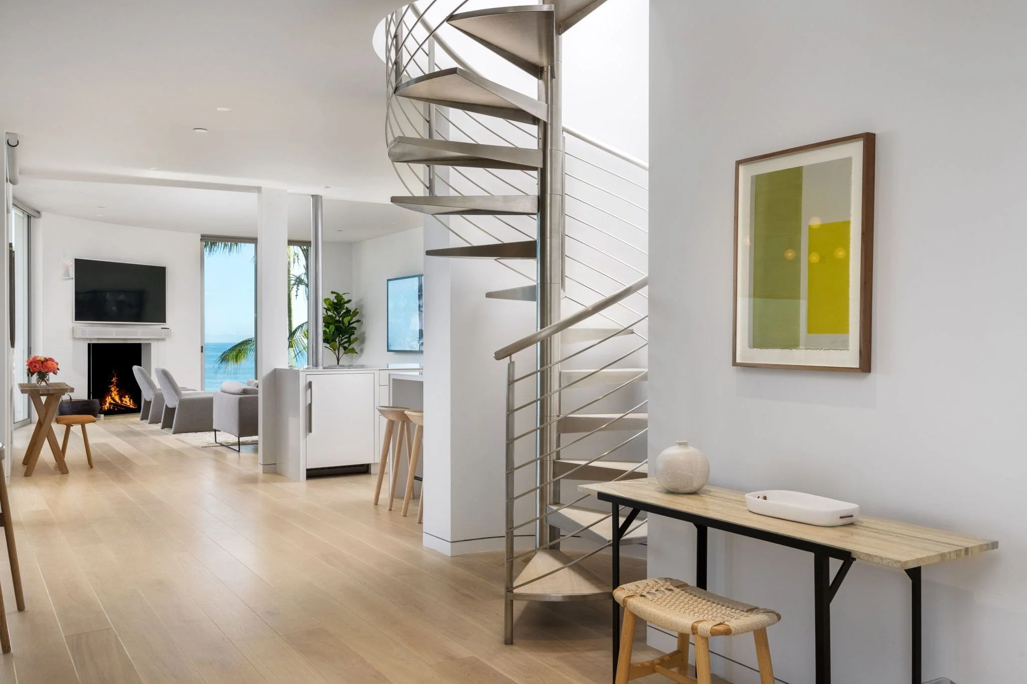 Open-concept living space with light wooden flooring, white walls, and a modern spiral staircase made of metal near a counter with a decorative vase. The living area has a fireplace, a wall-mounted television, and white chairs. Large windows reveal a