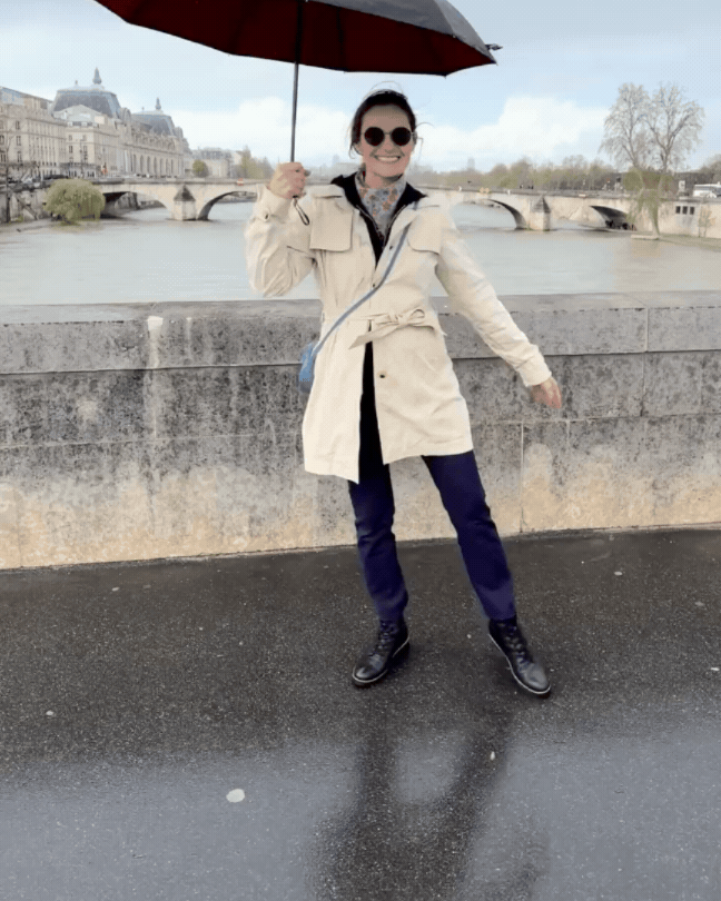 Woman standing on a wet sidewalk near a river, holding a black umbrella with a red underside, smiling, wearing sunglasses, a beige coat, dark pants, and black boots, with historical buildings and a bridge in the background.