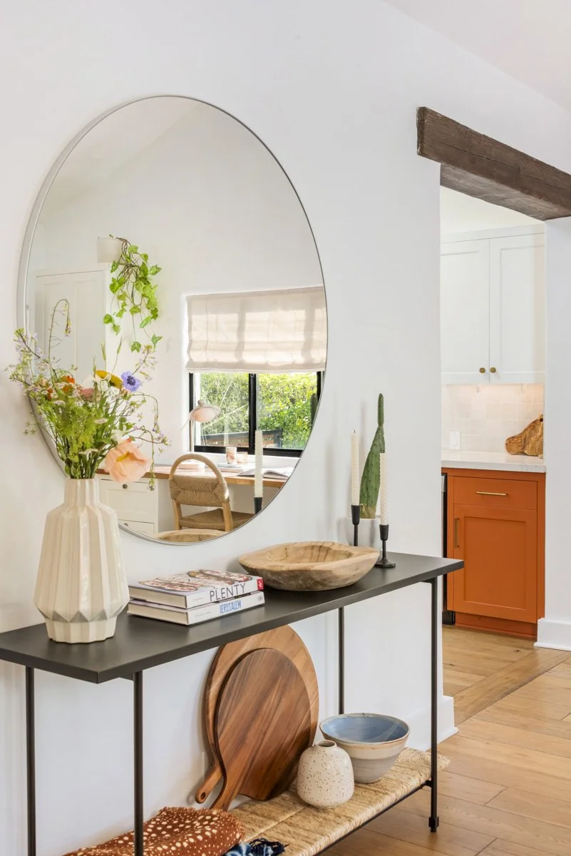 Interior view of a modern living space with a black console table decorated with a white vase holding flowers, books, a wooden bowl, candlesticks, and decorative bowls. A large oval mirror hangs above the table, reflecting a window with a view of gre