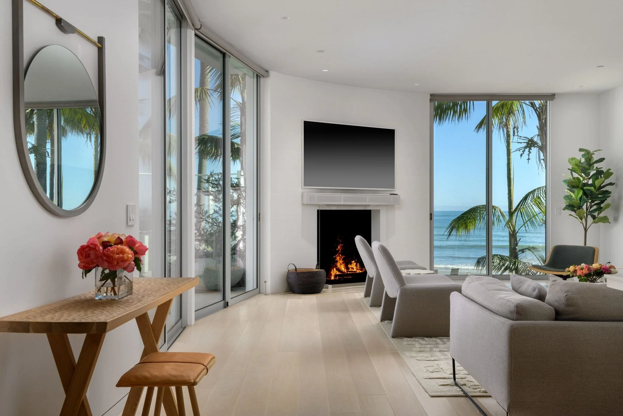 Modern living room with large glass windows and doors overlooking the ocean, featuring a fireplace, gray sofa, dining chairs, a side table with flowers, and a mirror on the wall.