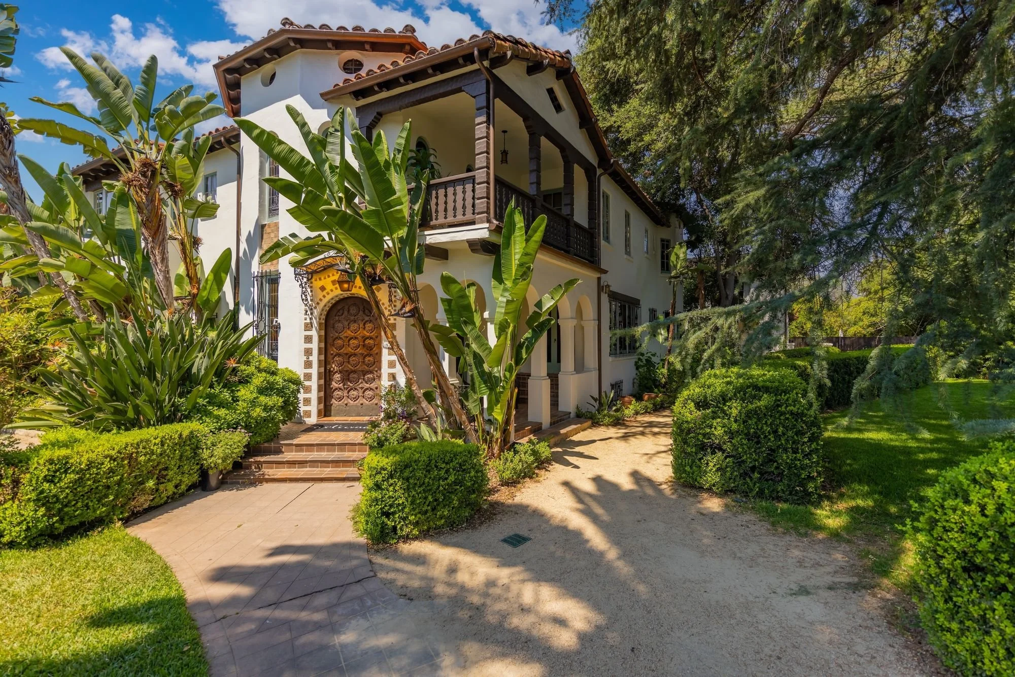 A white two-story house with a tiled roof, surrounded by lush green plants and tall trees, with a small step leading to the front door and a pathway in the garden.