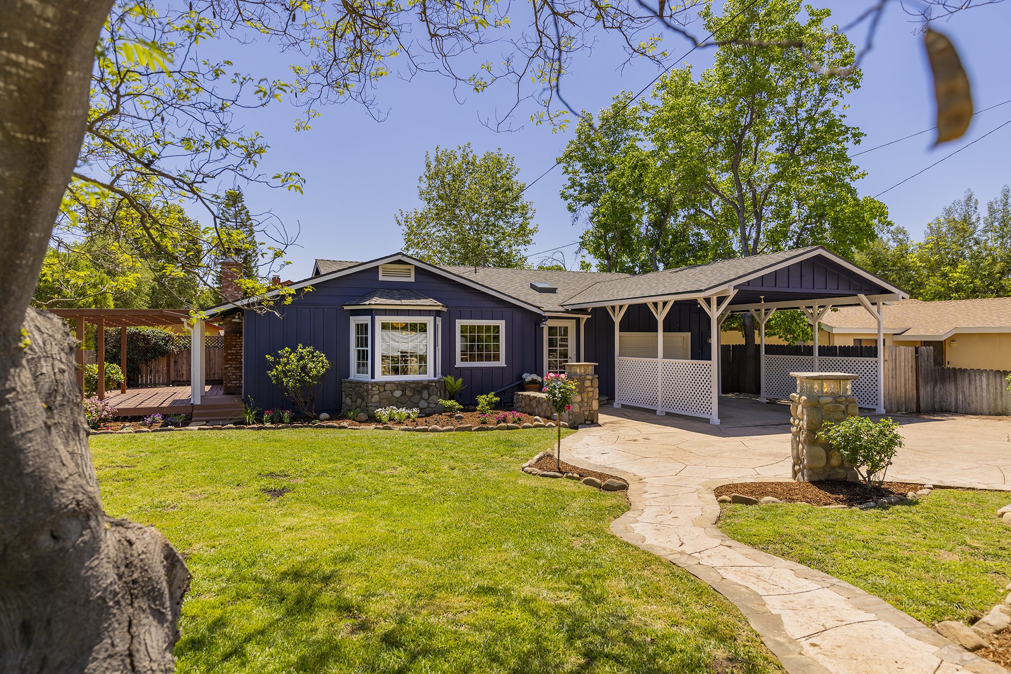 Front yard of a house with a well-maintained lawn, stone pathway, trees, a blue house with white-trimmed windows, a porch, and a white lattice fence.