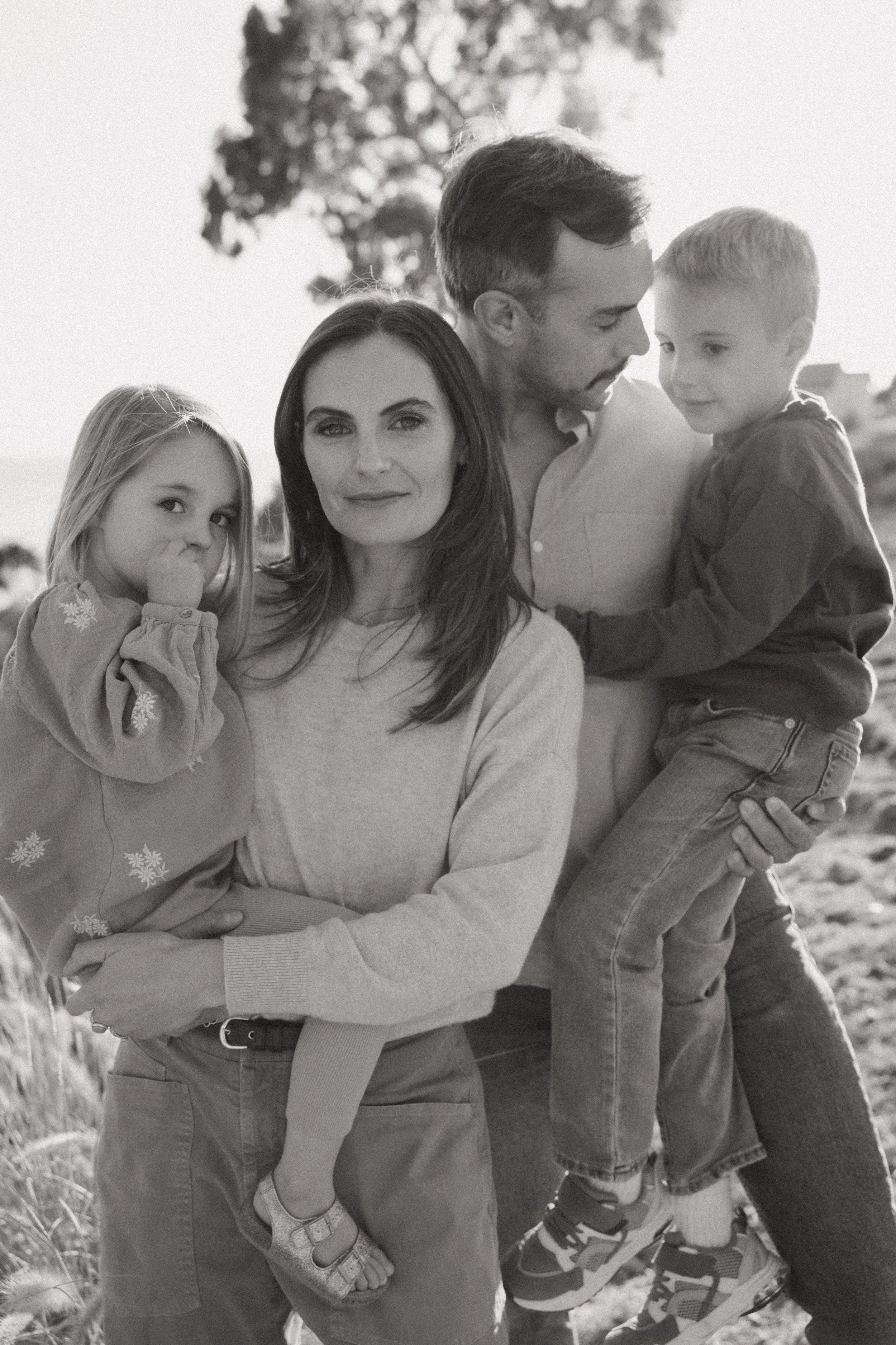 A black and white photo of a family outdoors. The mother is holding her young daughter on her hip, and the father is holding his young son. They are all looking happy and close together, with trees in the background.