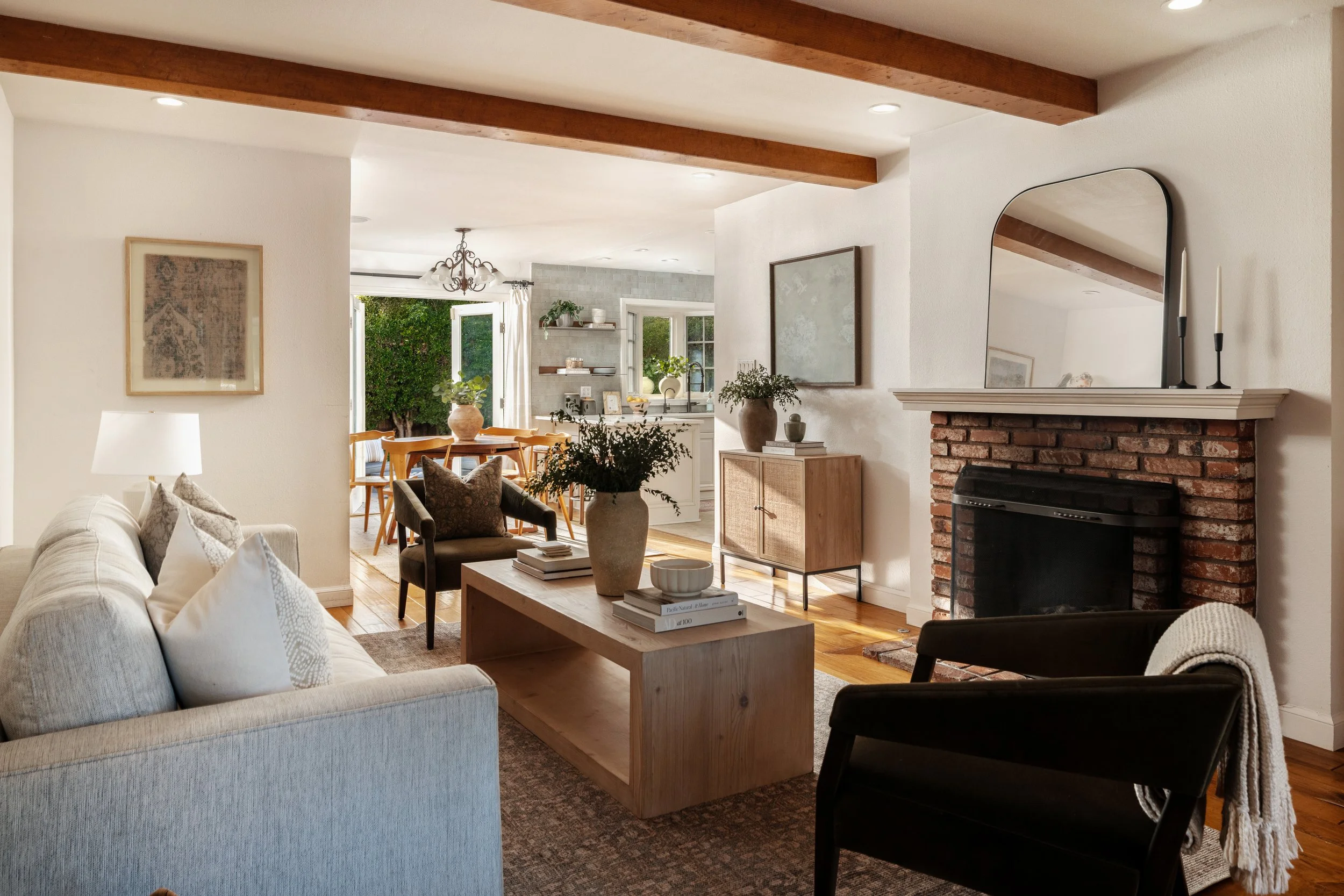 Living room with white sofa, dark chairs, wooden coffee table, brick fireplace with mirror, and greenery visible through windows.