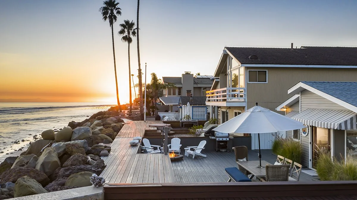 Beachfront houses with wooden deck and outdoor furniture at sunset, with palm trees and ocean in the background.