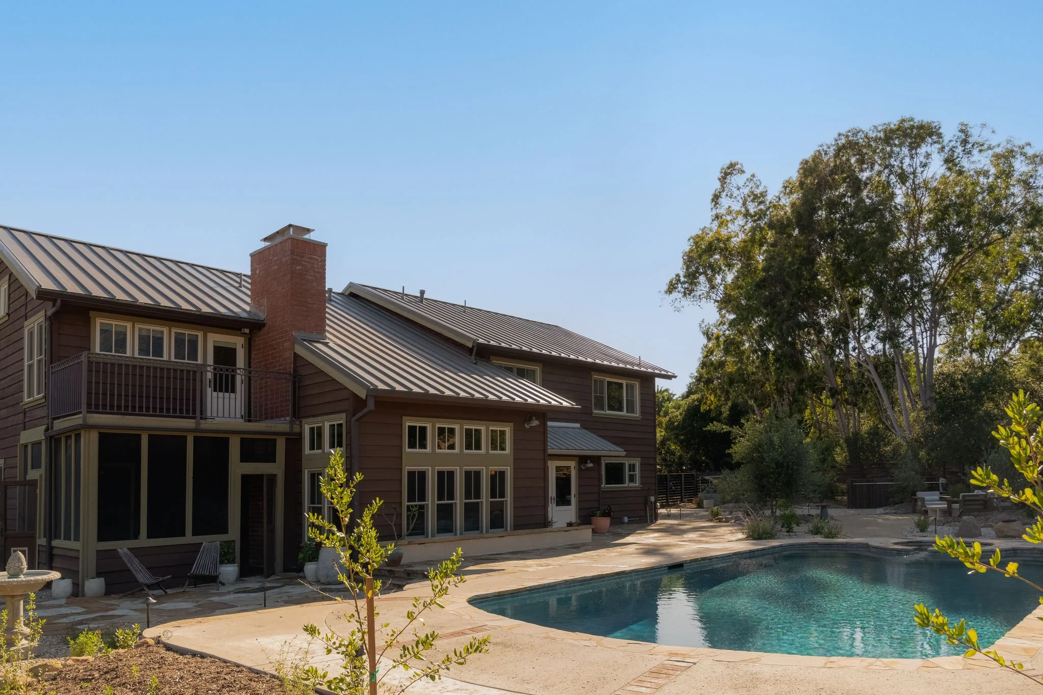 A backyard view featuring a multi-story house with a sloped metal roof, a brick chimney, large windows, and a small patio with some chairs, next to a swimming pool, surrounded by trees and plants, under a clear blue sky.