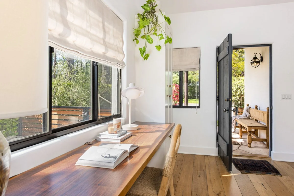 Interior view of a bright room with large windows, a wooden desk with books and a lamp, and a door leading to a porch with outdoor seating and greenery outside.