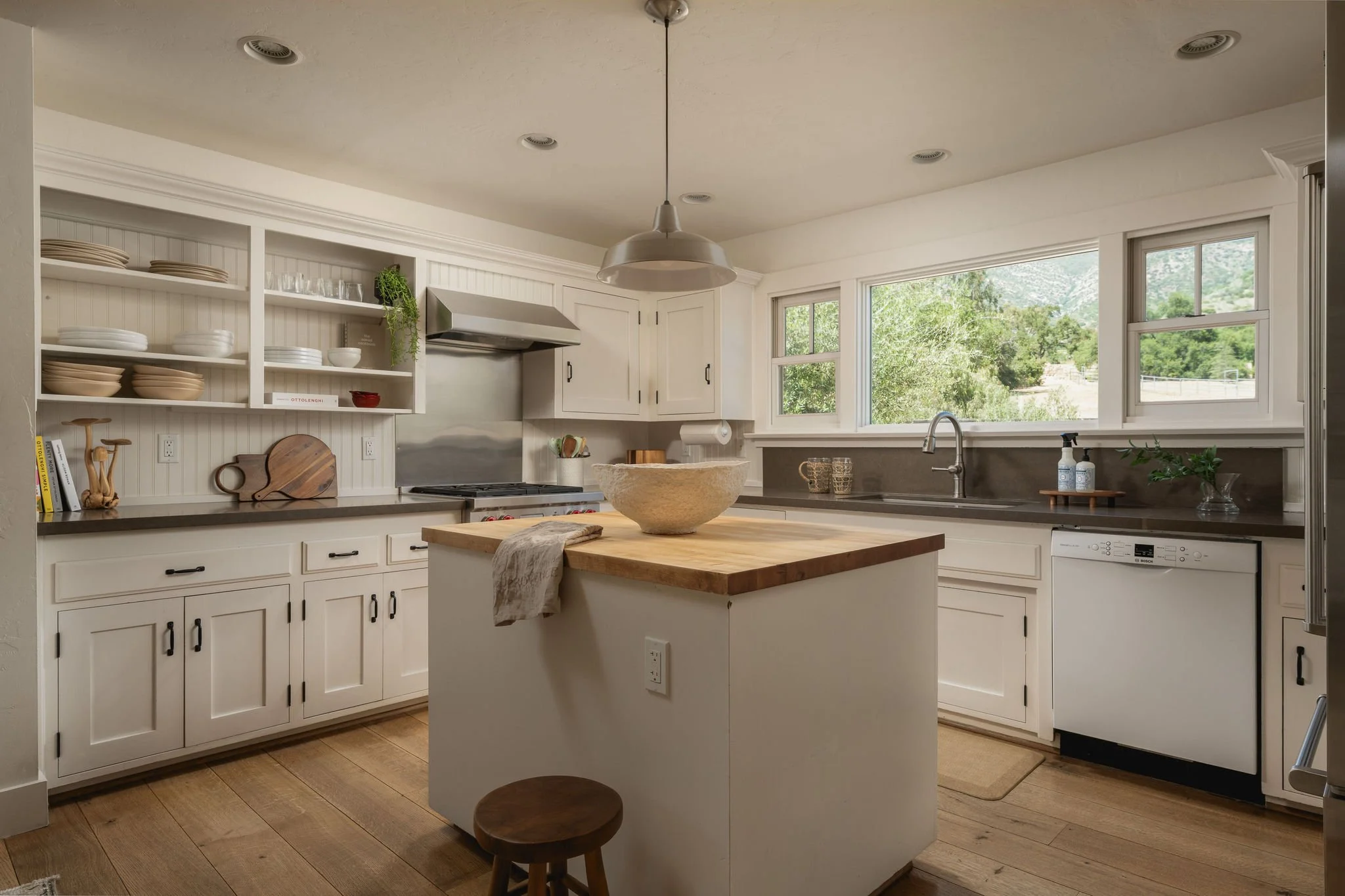 A bright kitchen with white cabinets, open shelves with dishes, a large window showing greenery outside, a kitchen island with a large stone bowl, and a small wooden stool in the foreground.