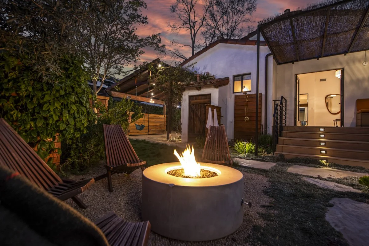 A backyard outdoor space at dusk with a fire pit, wooden chairs, a swing, and a white house with steps and lighting.