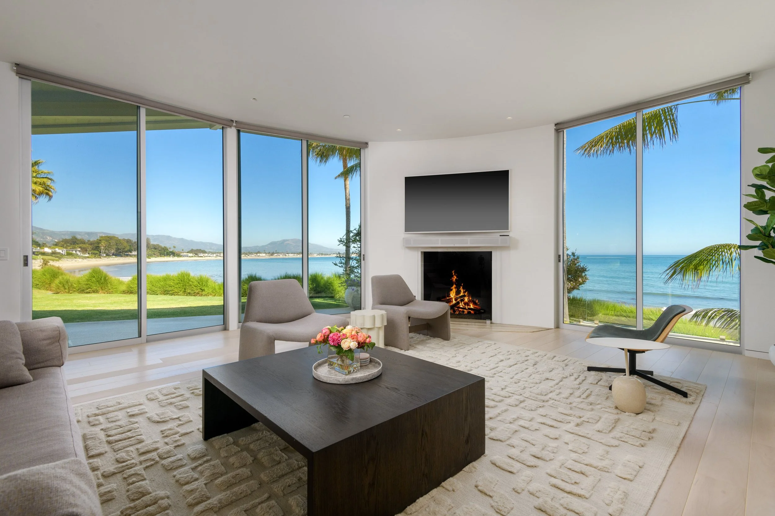 Living room with large floor-to-ceiling windows overlooking water and palm trees, modern furniture, a fireplace, and a flat-screen TV.
