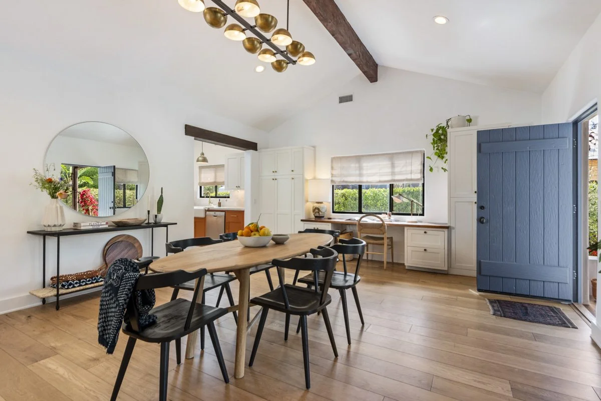 A dining area with a wooden table, black chairs, a sideboard with a large round mirror, and a view into the kitchen with white cabinets and black windows, all with a bright and airy atmosphere.