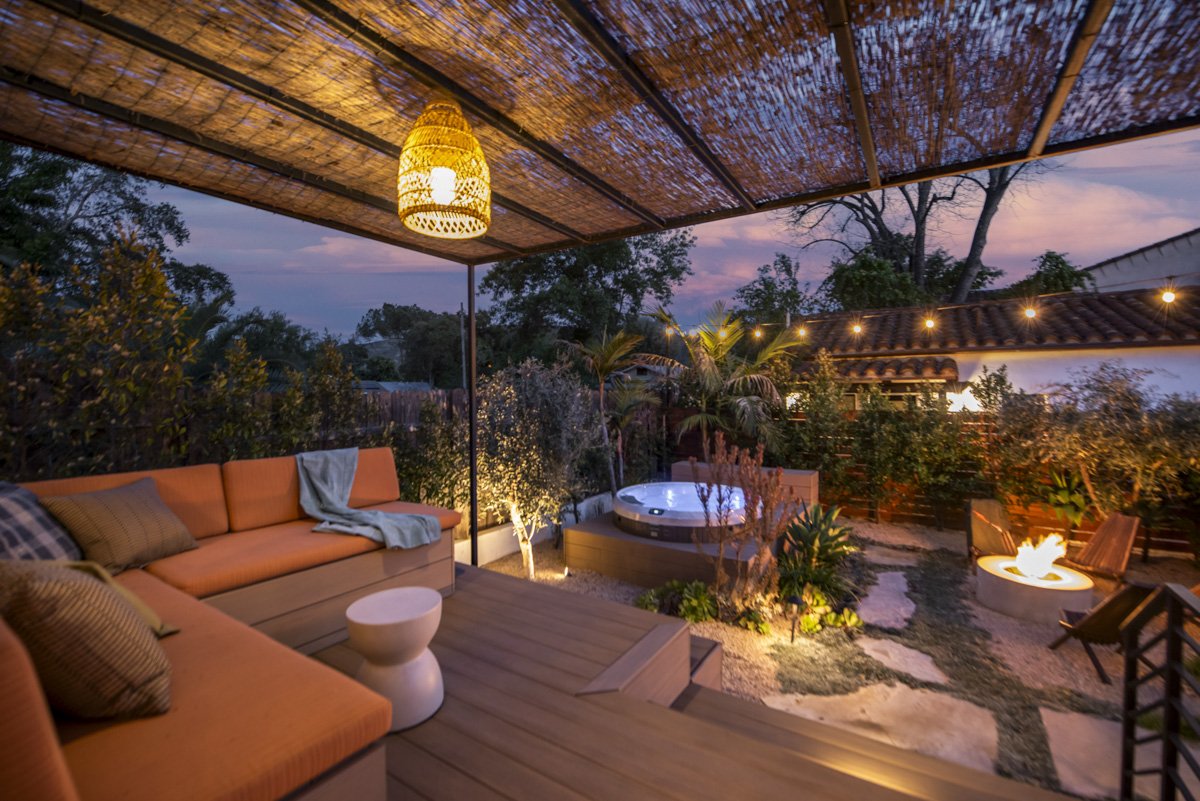 Outdoor patio with orange cushioned seating, a small white side table, a hot tub, a fire pit, string lights, trees, and a partly cloudy purple sunset sky.
