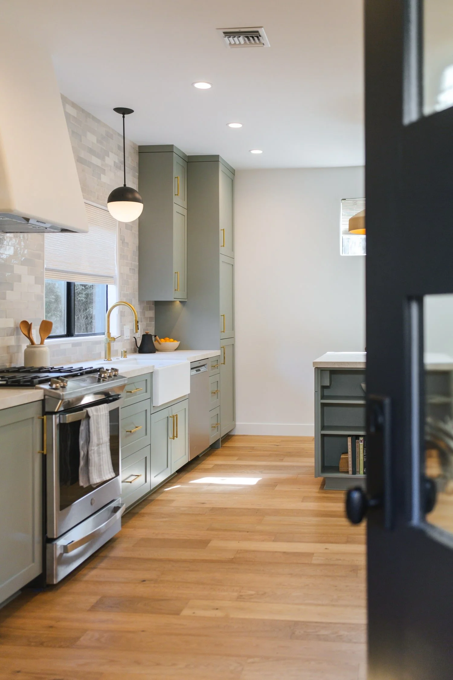 A modern kitchen with light green cabinets, a white countertop, a black pendant light, and hardwood floors. There is a window with blinds, a stovetop, and a small bookshelf on the right.