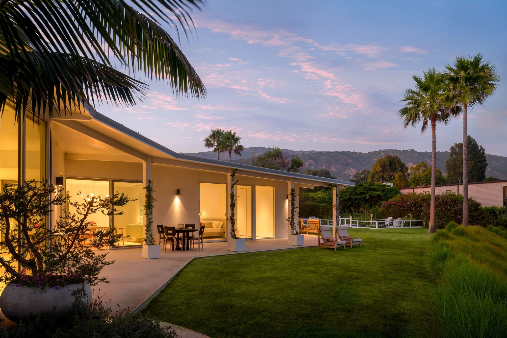Modern house with large windows, an outdoor patio, and a lush green lawn, with palm trees and mountains in the background during sunset.