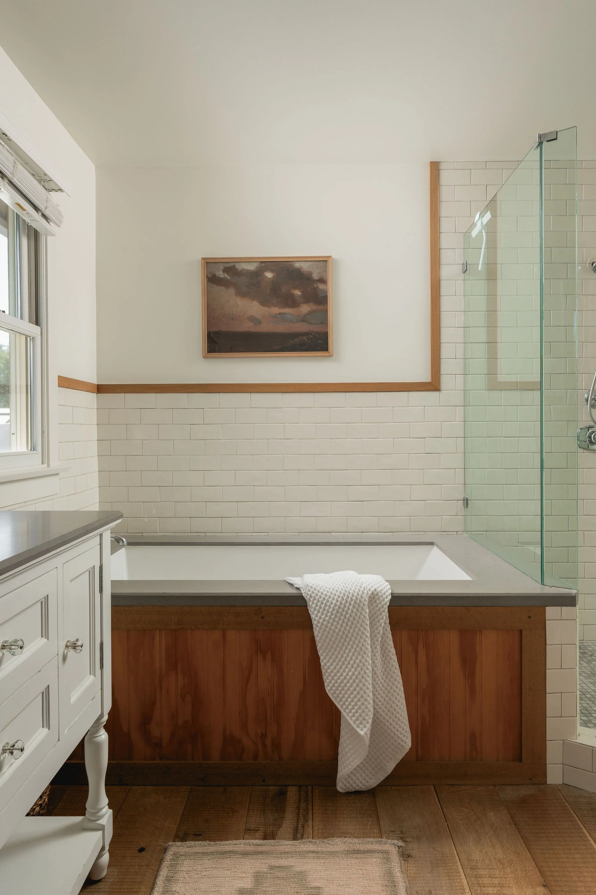 A bathroom with a bathtub and a wooden panel. There's a towel hanging over the bathtub's edge, a white dresser on the left side, a framed painting of a landscape with clouds on the wall, a window with blinds, and part of a glass shower enclosure to t